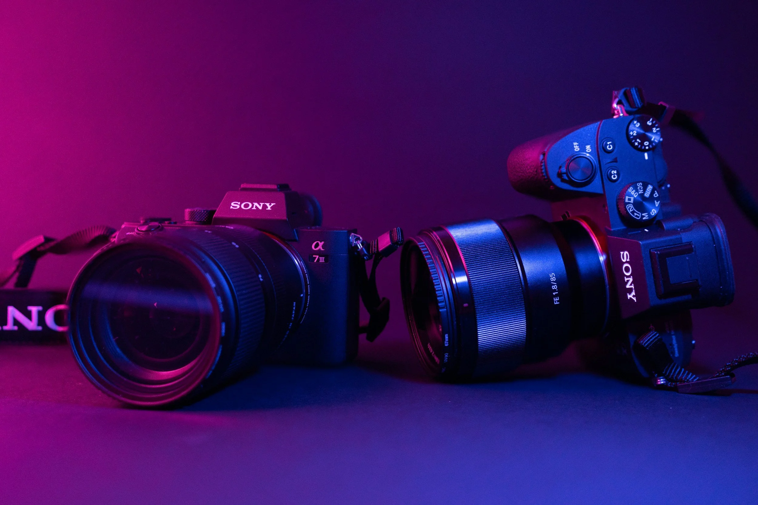 Close-up of two Sony cameras with lenses, illuminated with purple and pink lighting, placed on a dark surface.