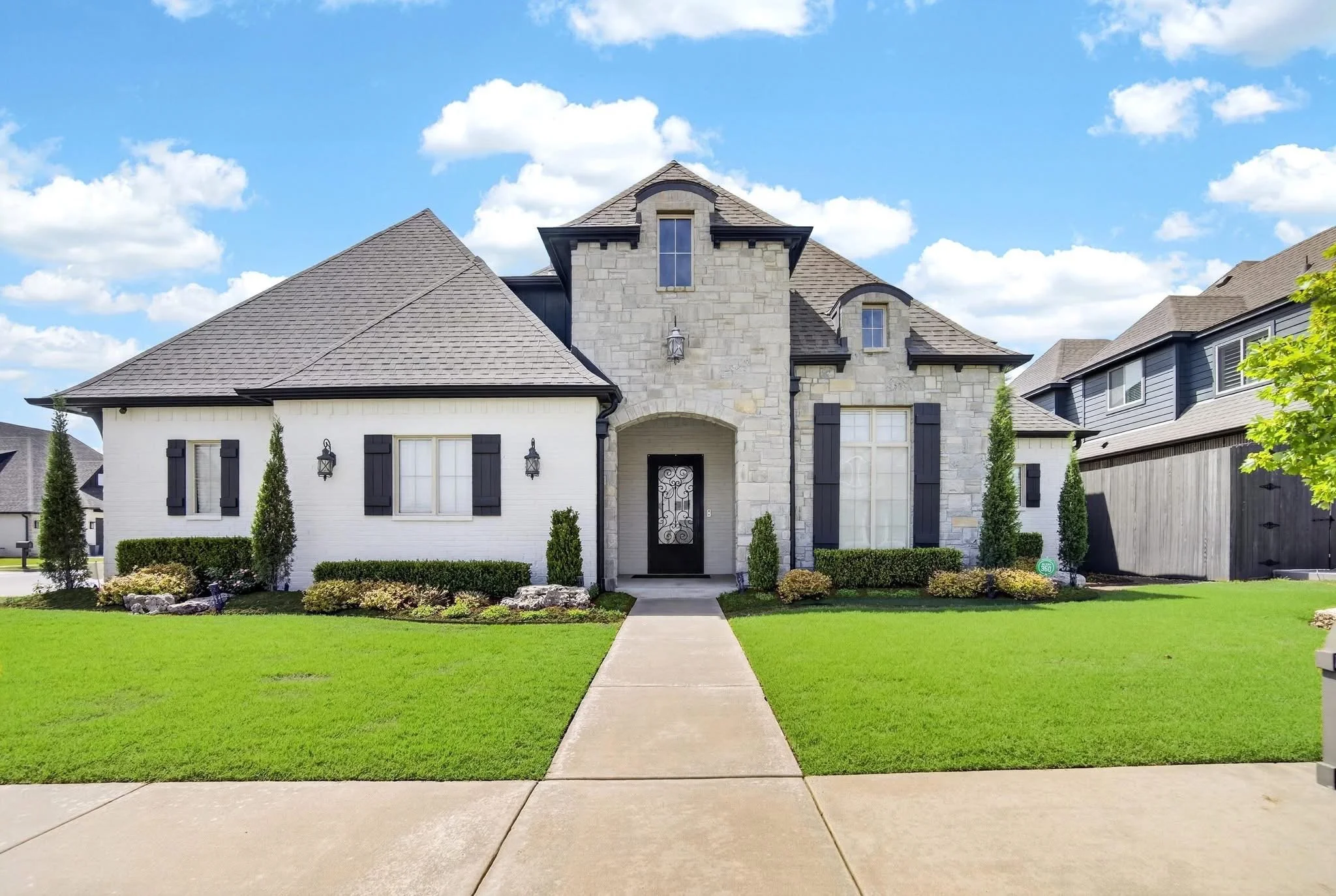 Front view of a two-story house with a stone and white exterior, black shutters, and a well-maintained front yard with a concrete walkway, green grass, and small shrubs, under a partly cloudy sky.