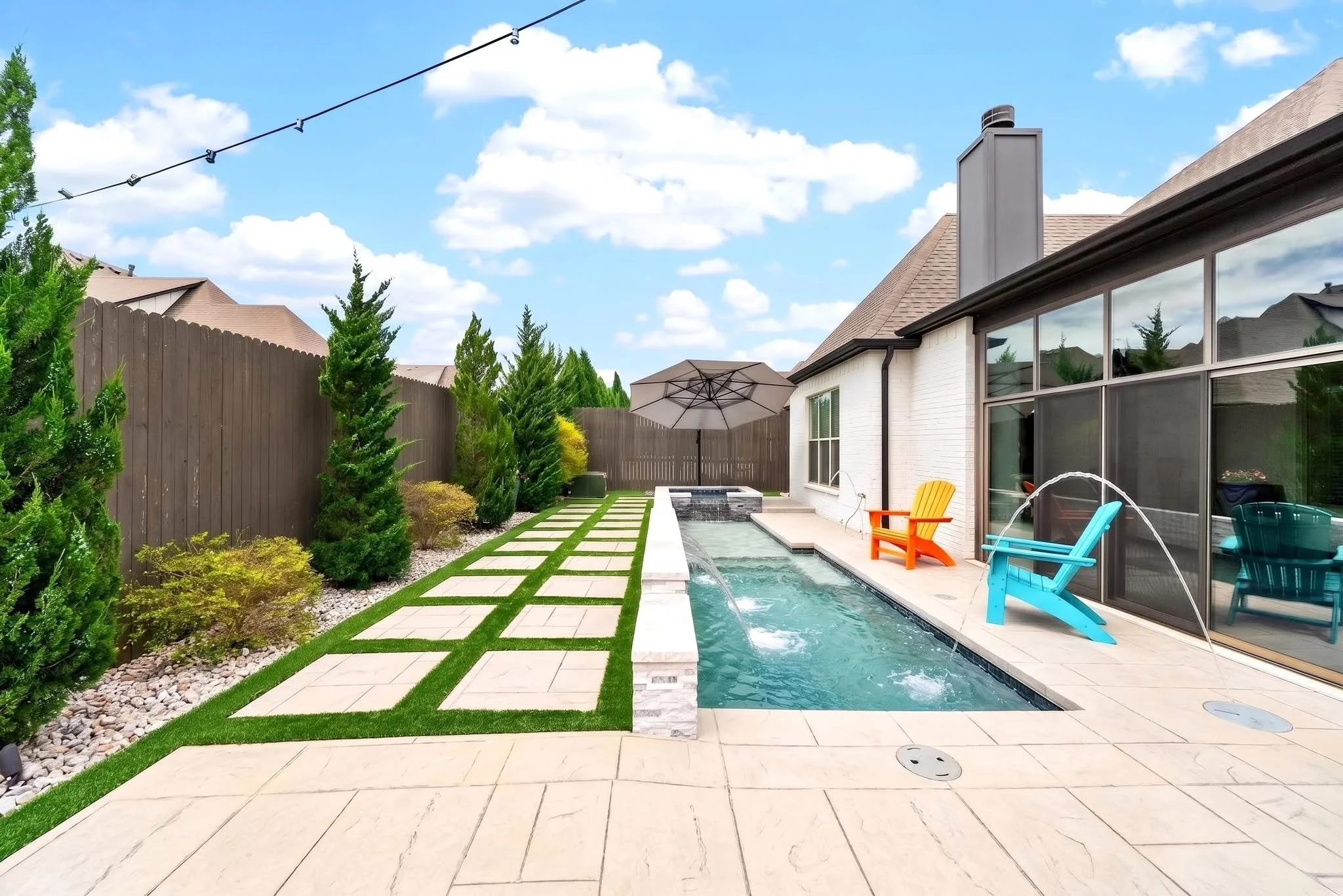 Backyard with a swimming pool, outdoor seating, potted plants, a concrete pathway, and a wooden fence with trees and a blue sky