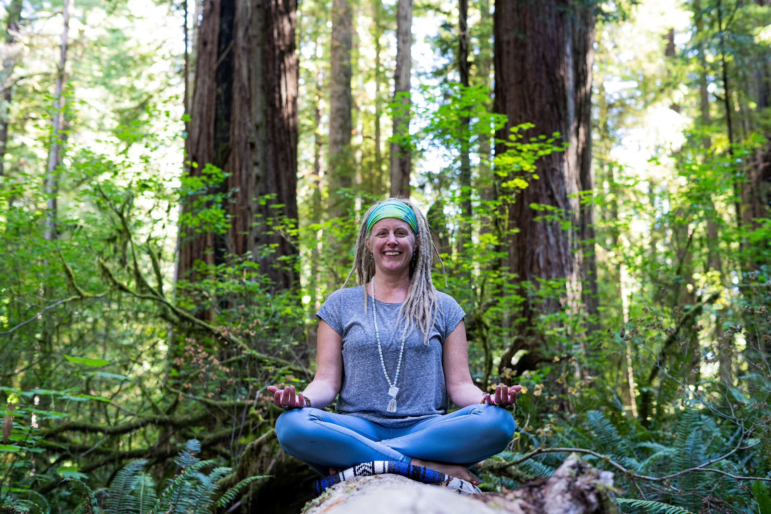 Woman with dreadlocks sitting in a cross-legged meditation pose on a fallen log in a lush green forest, smiling and wearing a gray t-shirt, colorful headband, and beaded necklace.