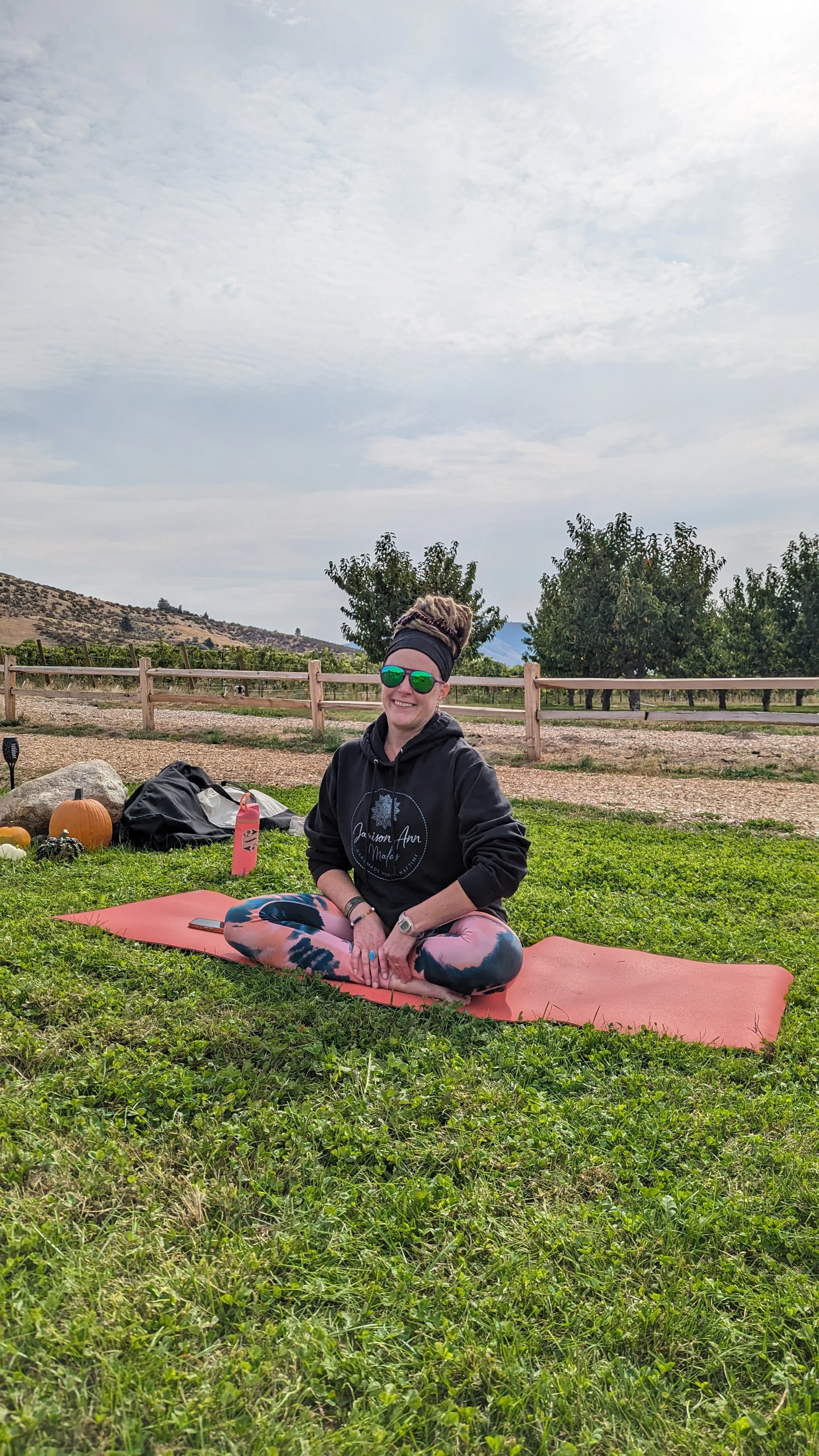 Woman sitting cross-legged on a pink yoga mat outdoors, wearing sunglasses, a black hoodie, and colorful leggings, with trees and a mountain in the background.