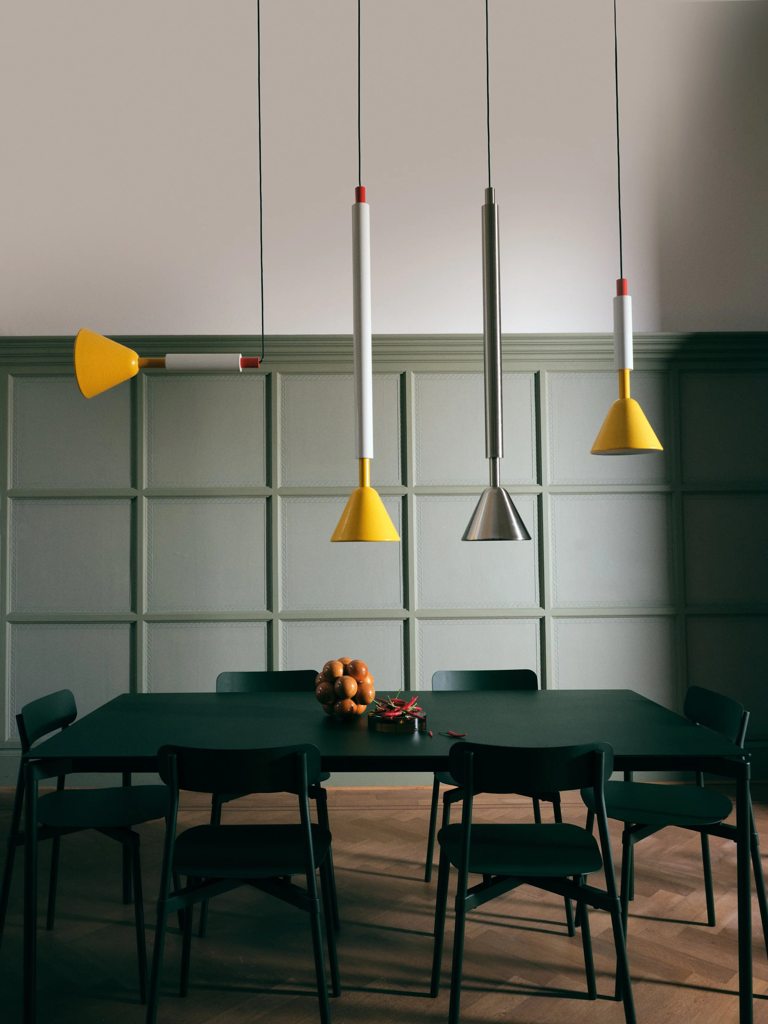 Salle à manger moderne avec un grand table noir, six chaises noires, un mur vert pastel avec panneaux en bois, trois suspensions de lumières modernes au plafond, un meuble au centre de la table avec quelques objets décoratifs.