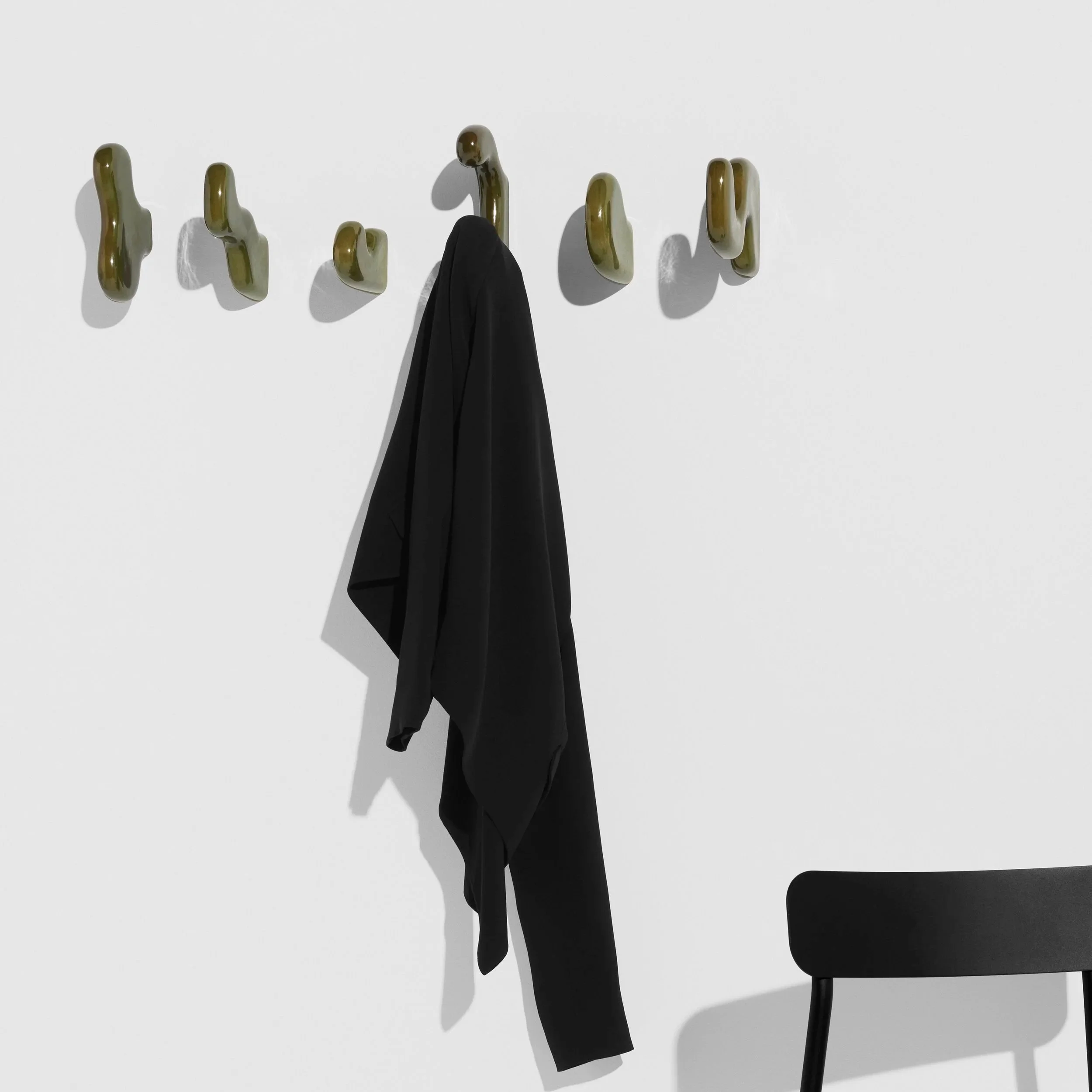 Une veste noire suspendue à un crochet doré, sur un mur blanc avec plusieurs crochets dorés, et une chaise noire à droite.