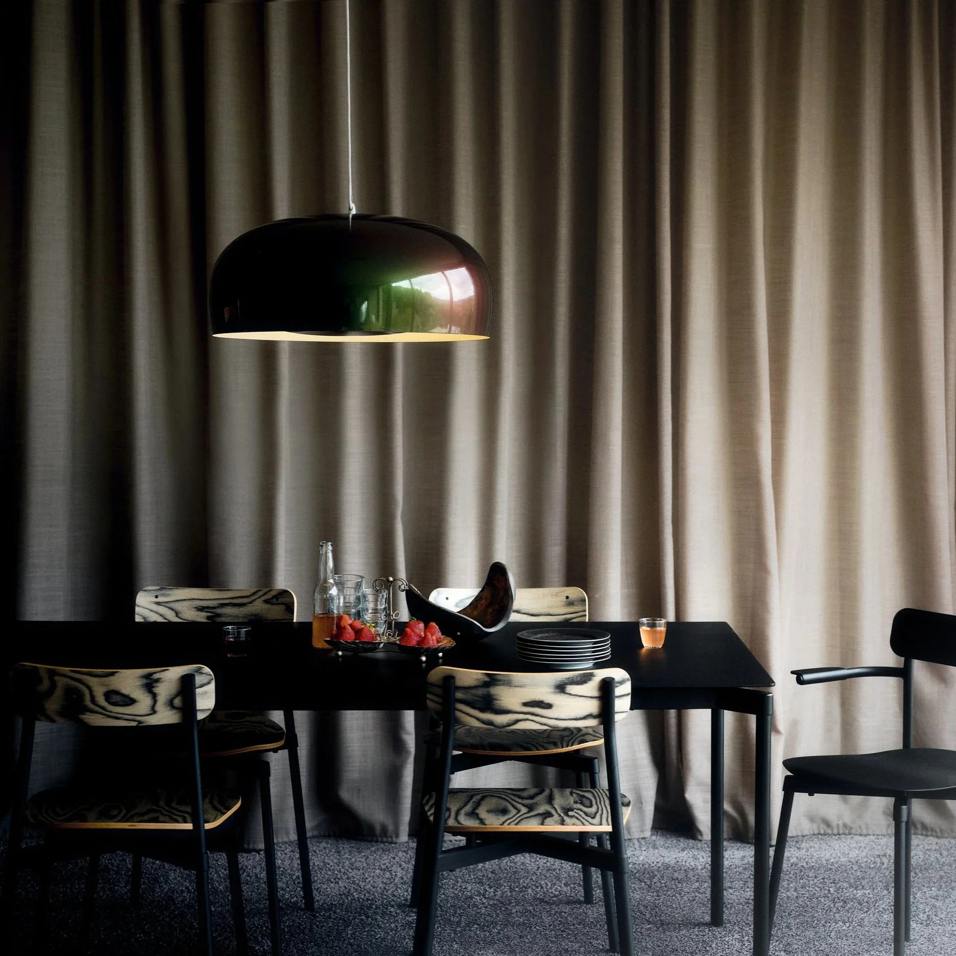 Une table à manger noire avec des chaises en bois grainé, décorée de fruits, de bouteilles, de verres, et de vaisselle, dans une pièce aux rideaux beige