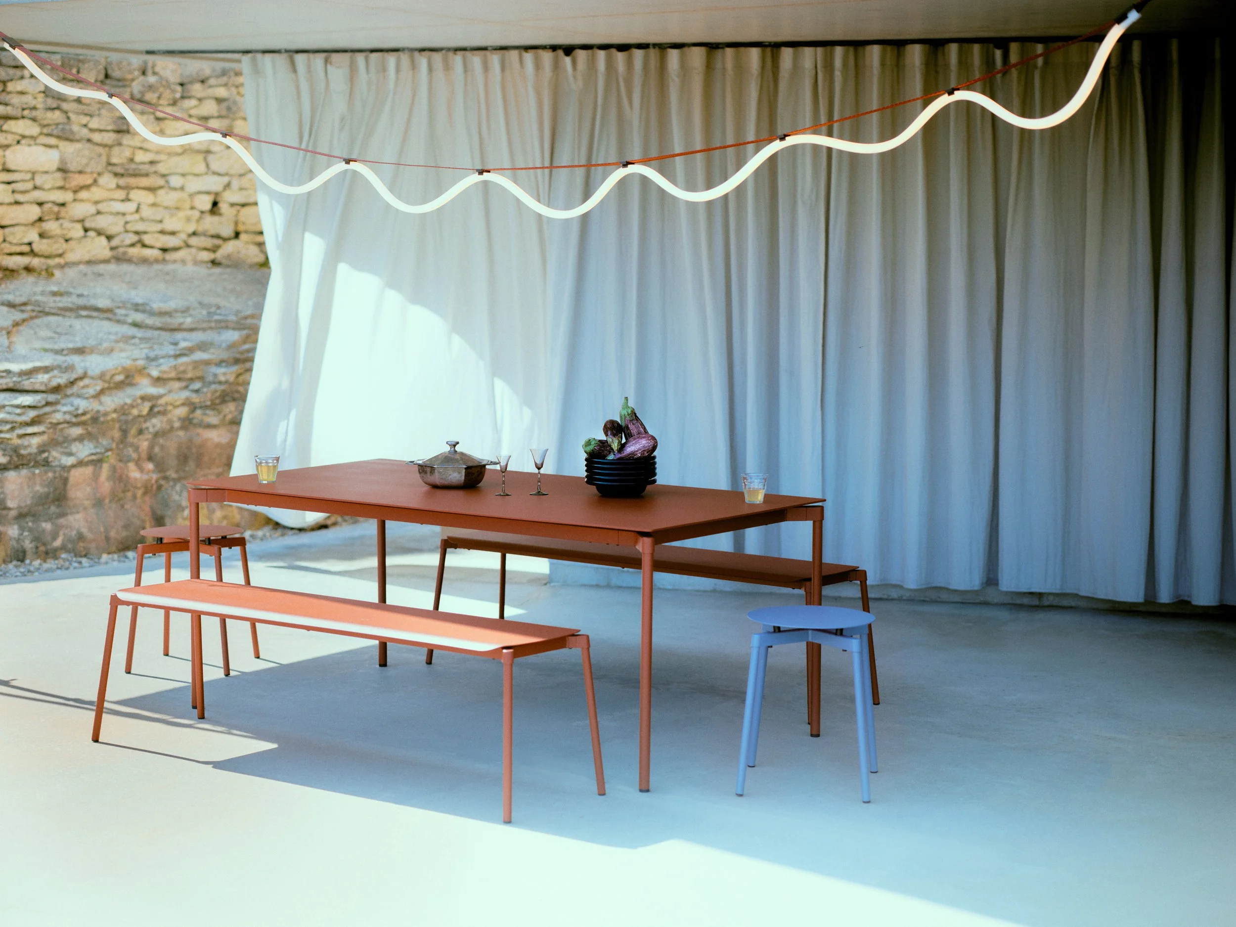 Table orange avec bancs et tabourets, décor simple sous une tente avec rideaux blancs, décorations avec des fruits, et deux verres devant.