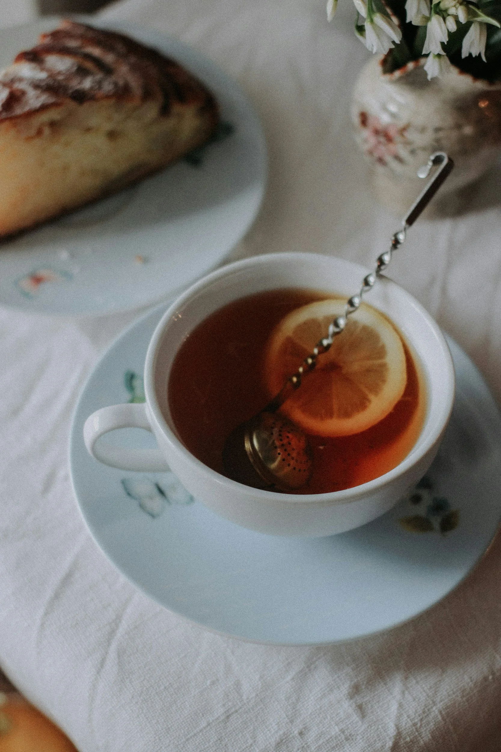 A white tea cup with a lemon slice and tea infuser on top, placed on a saucer with floral design. In the background, there is a slice of cake on a plate and a vase with flowers on a white tablecloth.