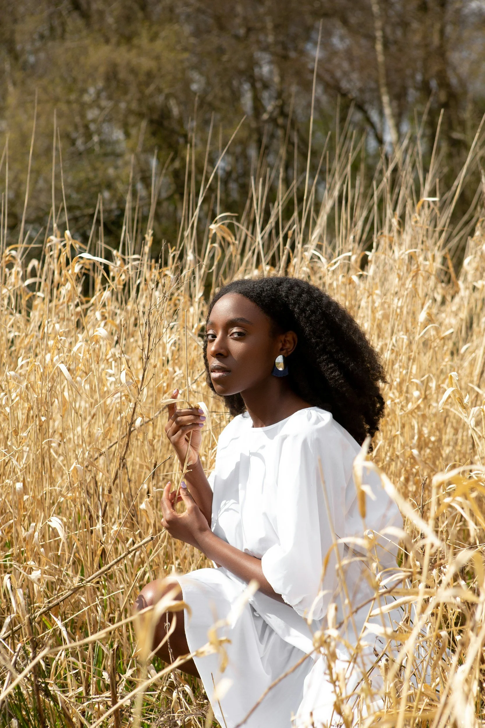 A woman with dark skin and curly hair is sitting in a field of tall, dry grass, wearing a white dress. She is looking at the camera and holding a piece of grass.