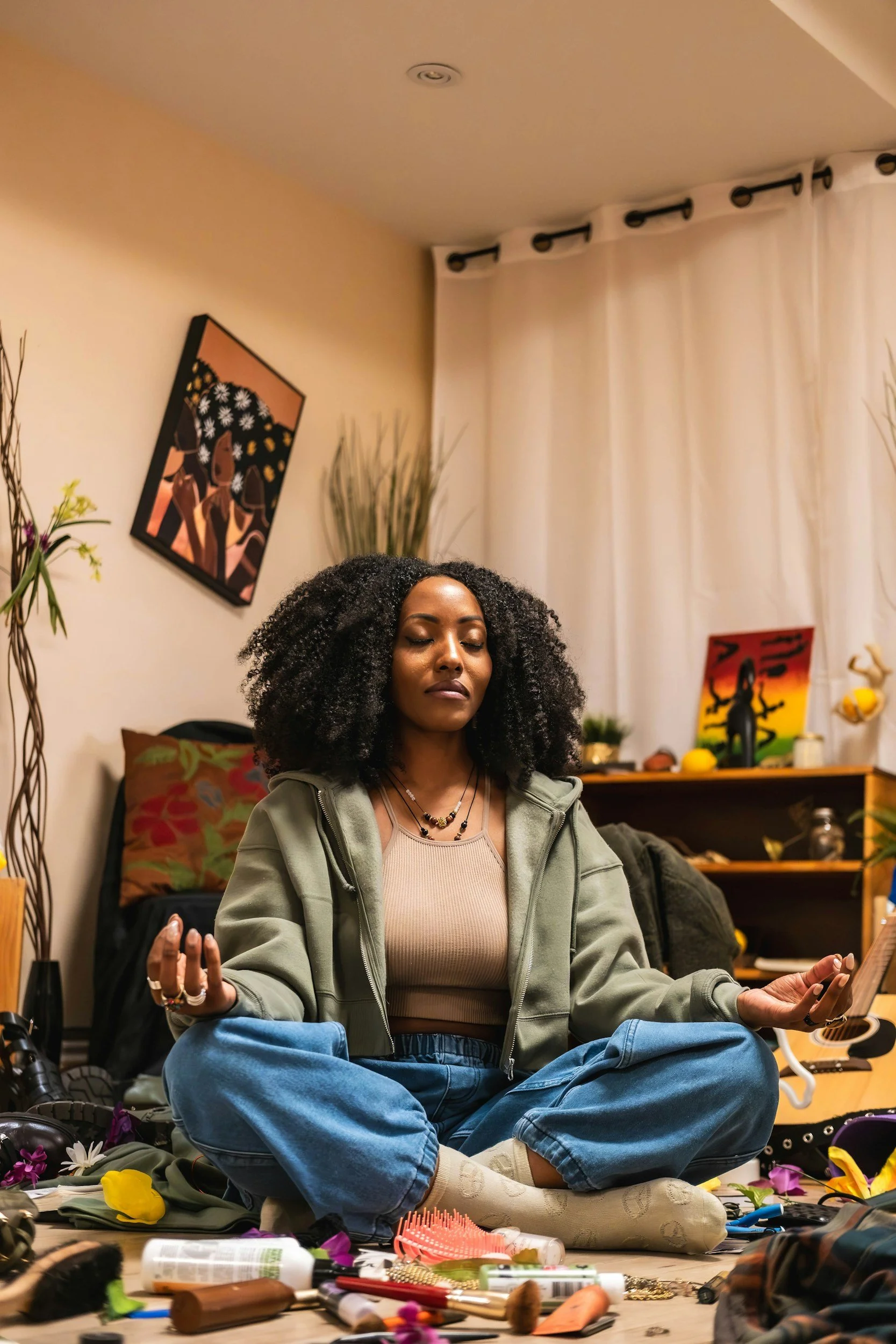 A woman with curly black hair sitting cross-legged on the floor in a room, meditating with closed eyes, surrounded by various objects such as flowers, makeup, and a guitar.