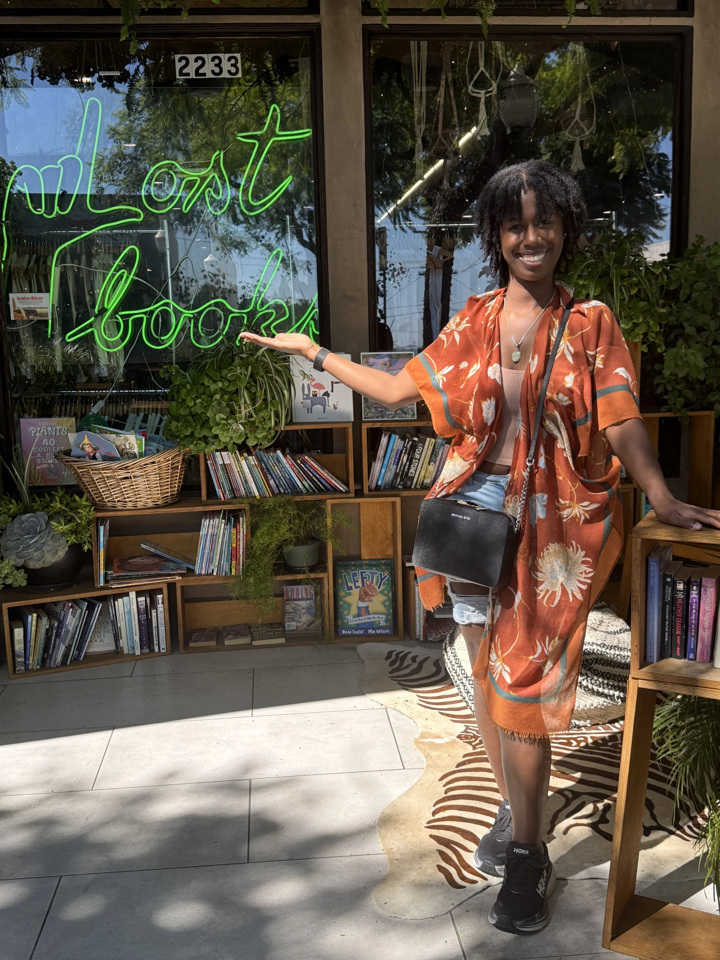 A woman with curly hair smiling and wearing a floral orange shirt with a necklace, standing in front of a bookstore window with a neon sign that says 'wildest books.' She is pointing at the sign with her left hand and holding a small black purse in her right hand.