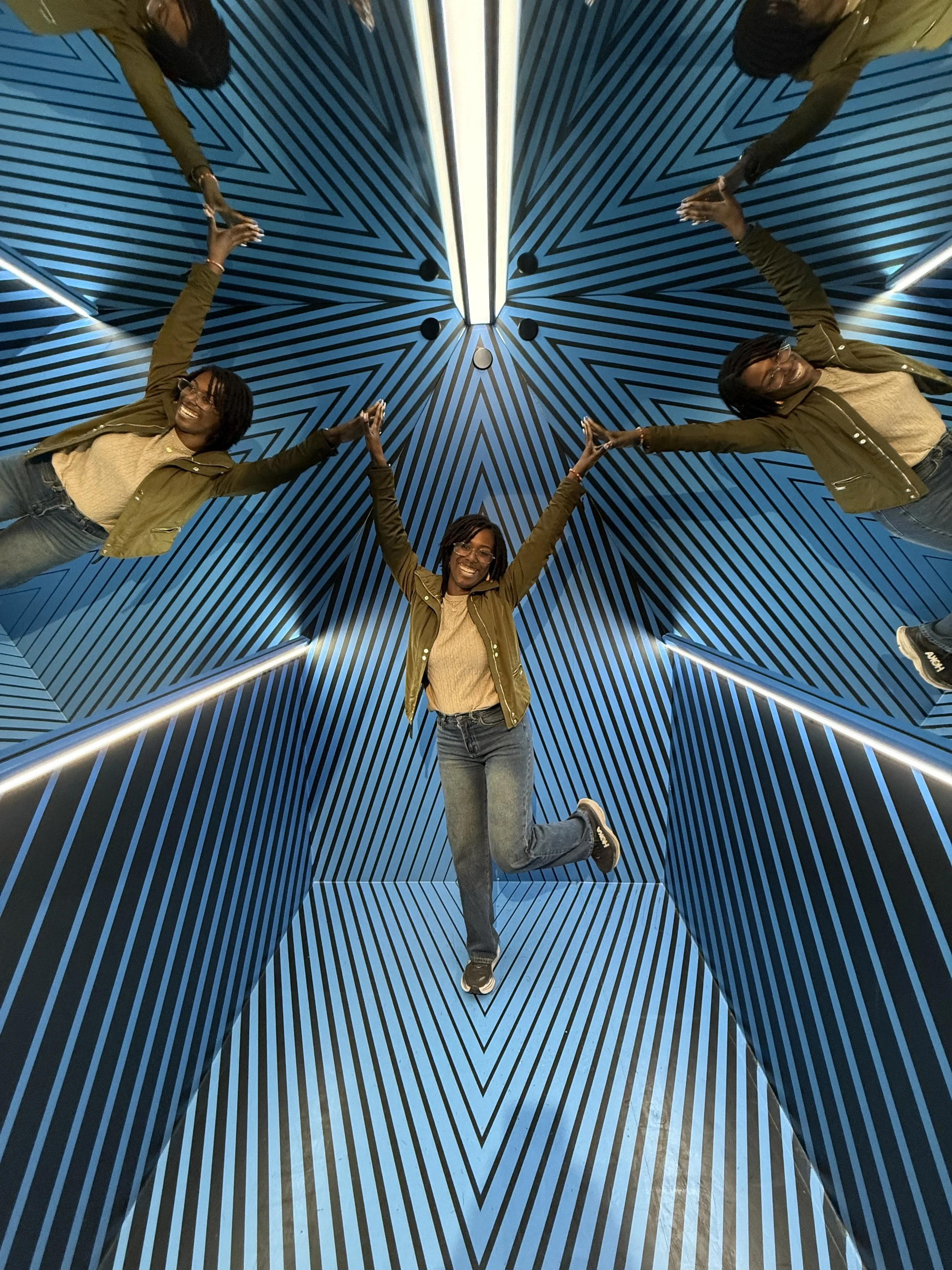 A woman with glasses in a brown jacket and blue jeans poses in an optical illusion room with blue and black striped walls forming a star pattern on the ceiling, and white lights along the edges. She is smiling, with one leg lifted, and her arms raised and joined with reflections creating the illusion of a larger figure.