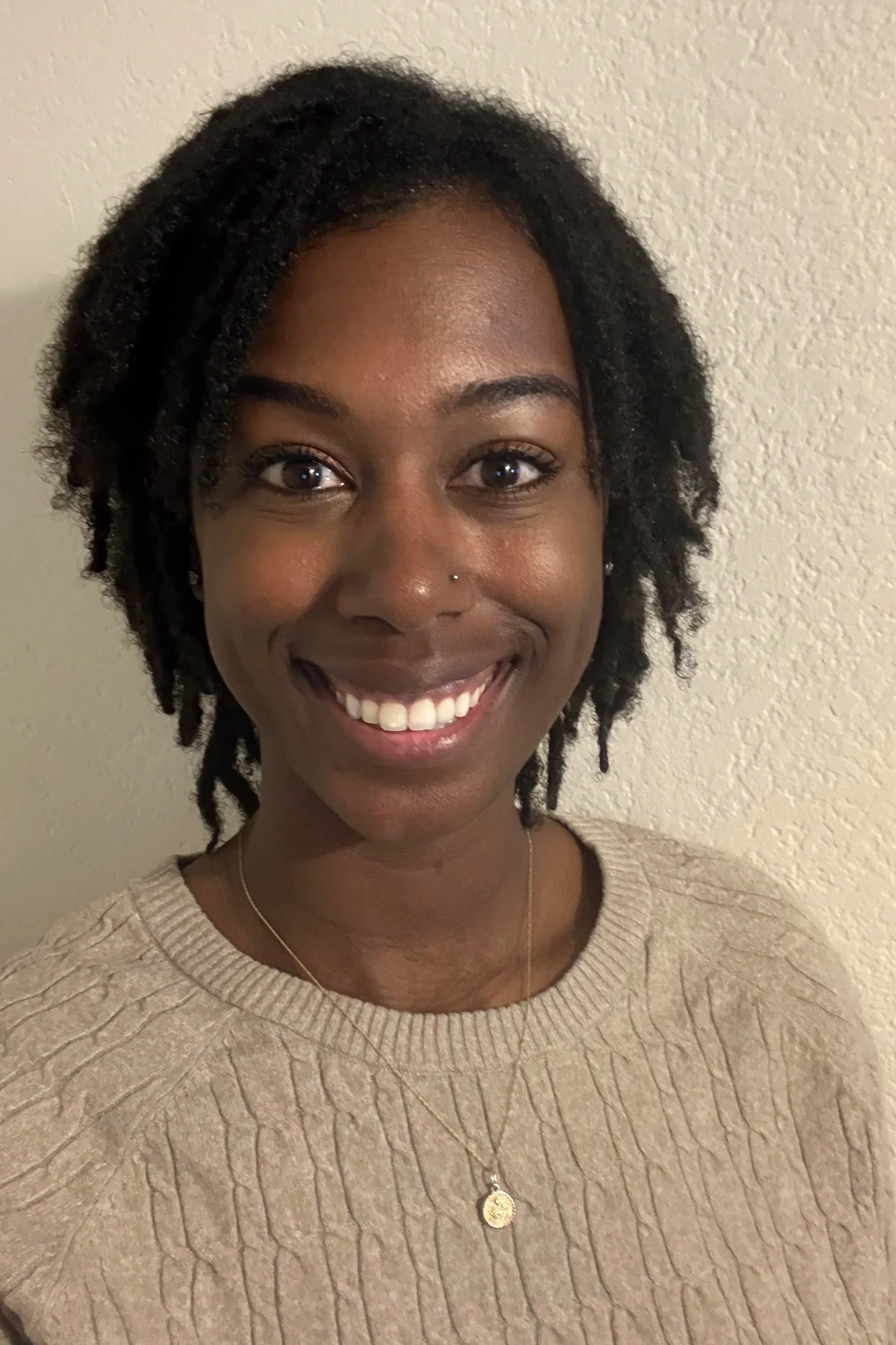 Close-up photograph of a smiling woman with short, curly black hair, wearing a beige sweater and a necklace, standing against a neutral textured wall.