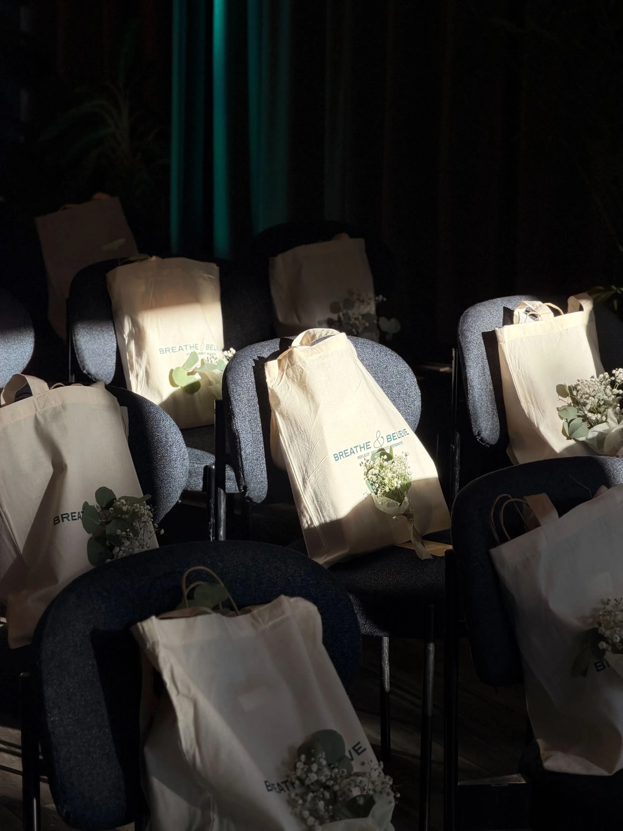 Multiple black chairs with cream-colored tote bags and small floral arrangements on the seats, set in a dark room with teal curtains.