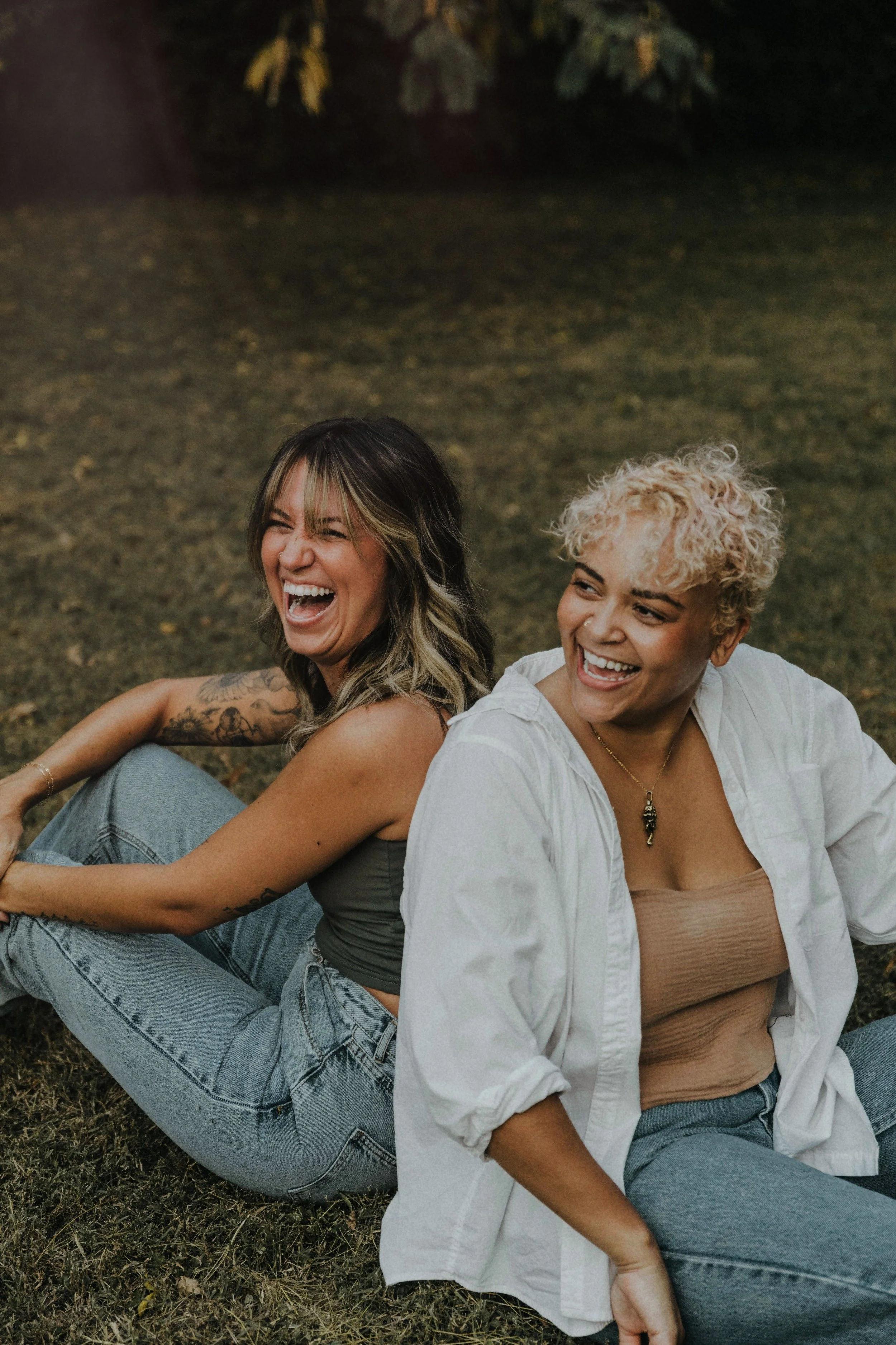 Two women laughing and smiling, sitting on the grass in a park at sunset.
