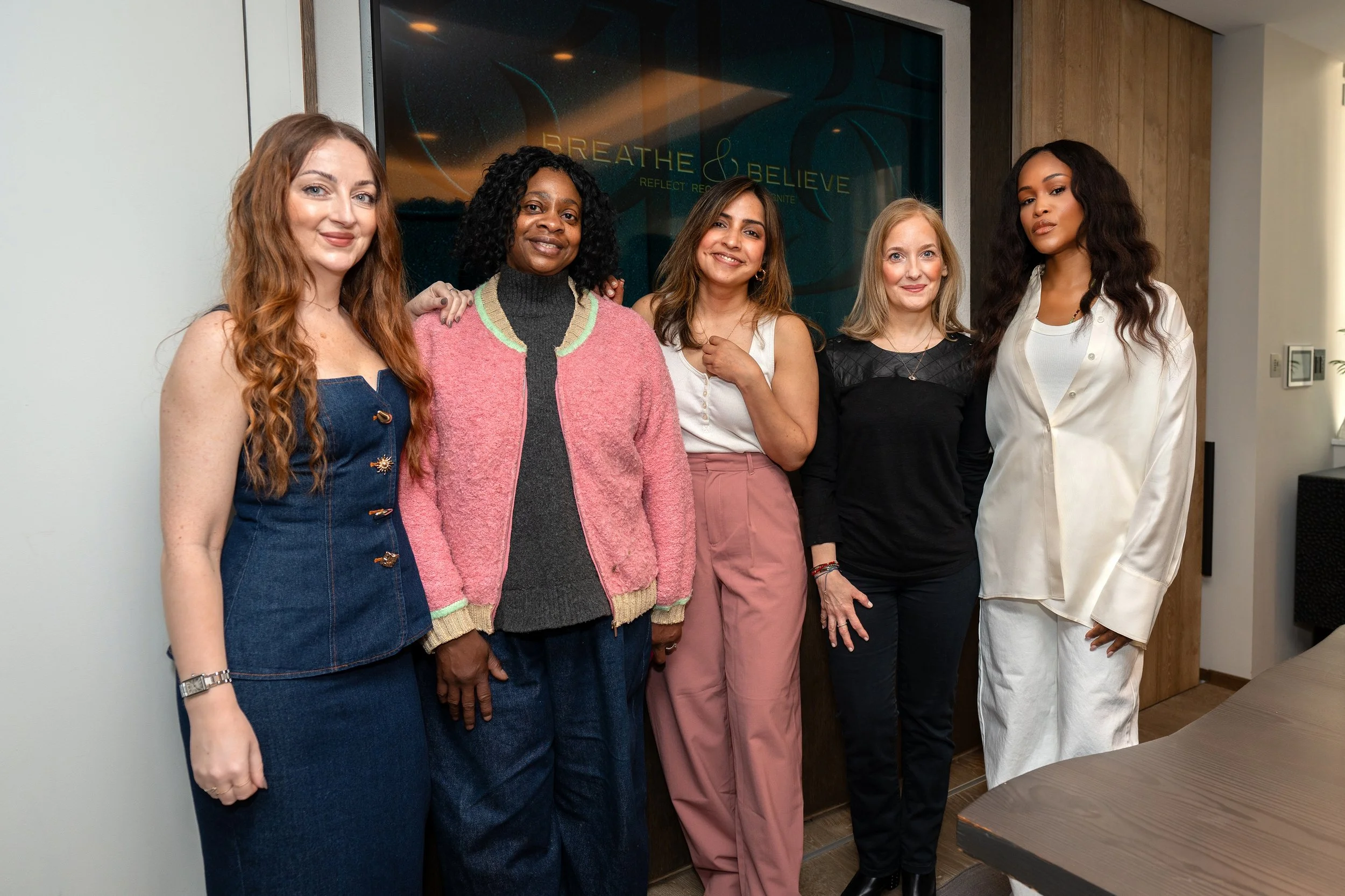 A diverse group of six women standing together indoors in front of a dark wall with a screen that has the words 'BREATHE & BELIEVE' on it. They are smiling and appear to be at a professional event or meeting.