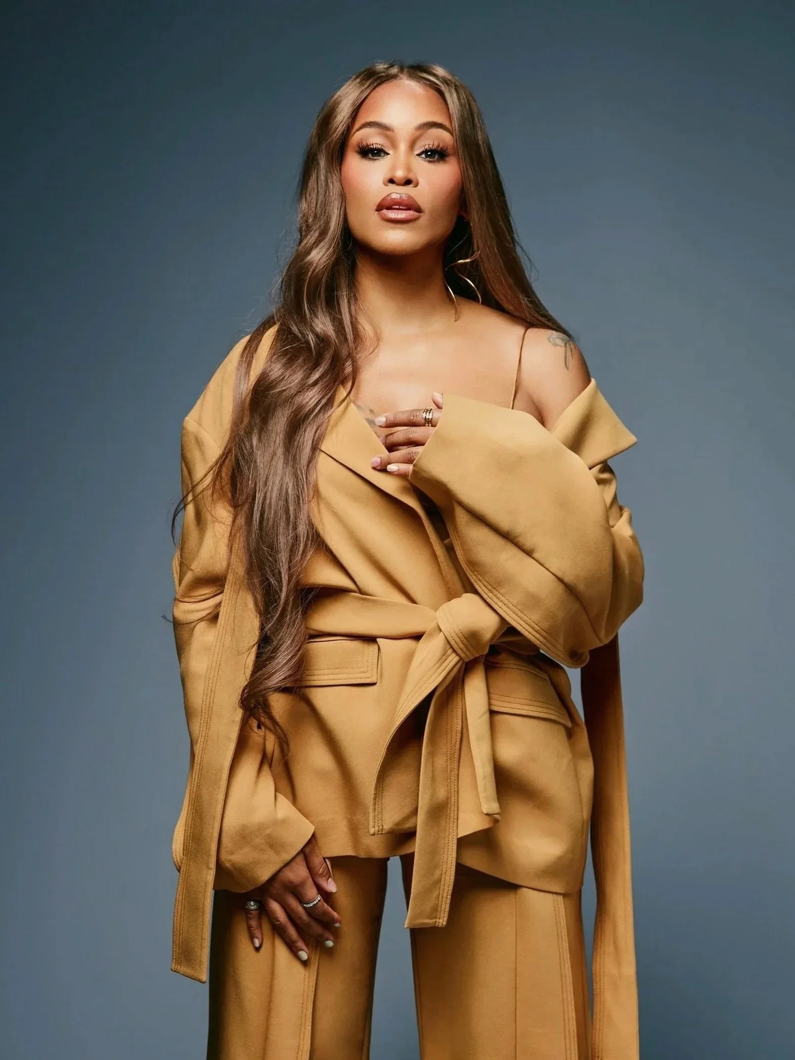 A woman with long wavy hair and makeup, wearing a yellow suit with a jacket draped over her shoulders and tan pants, posing against a neutral background.