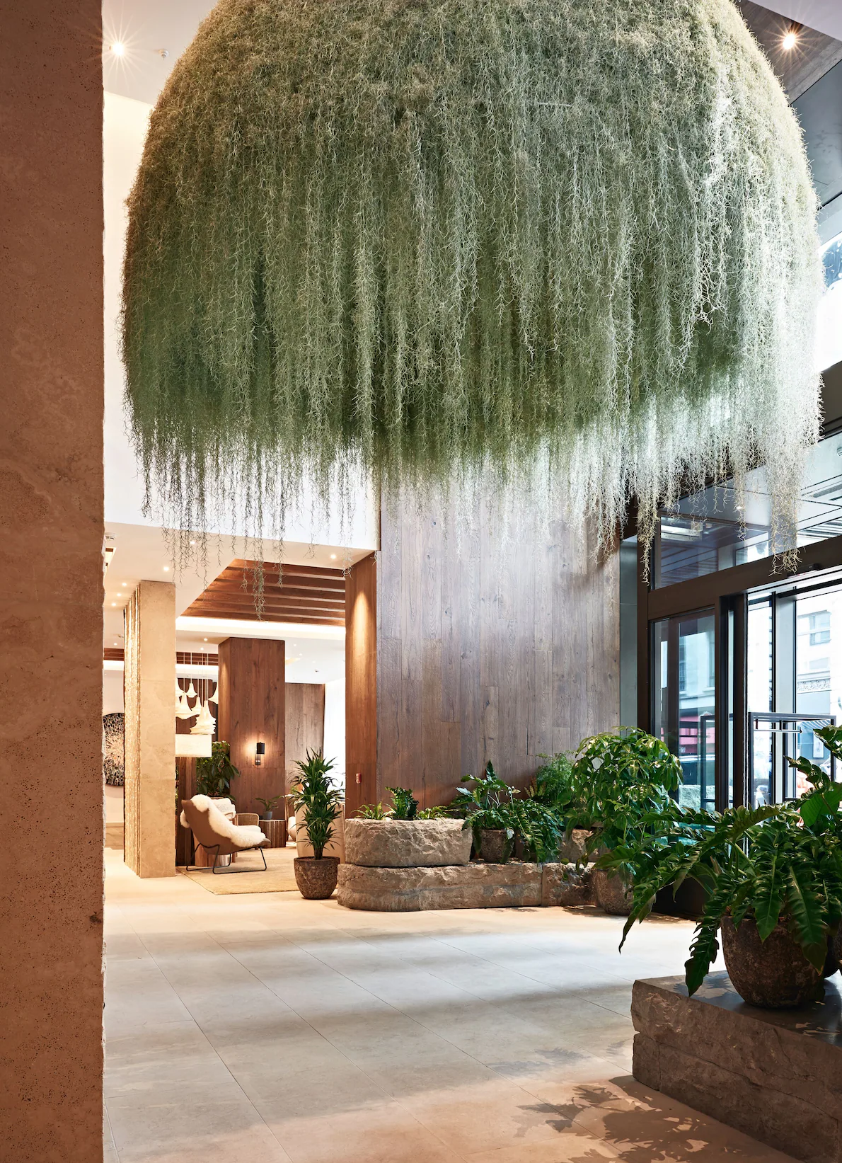Indoor hotel lobby with large hanging moss sculpture, potted plants, modern furniture, and large glass windows.
