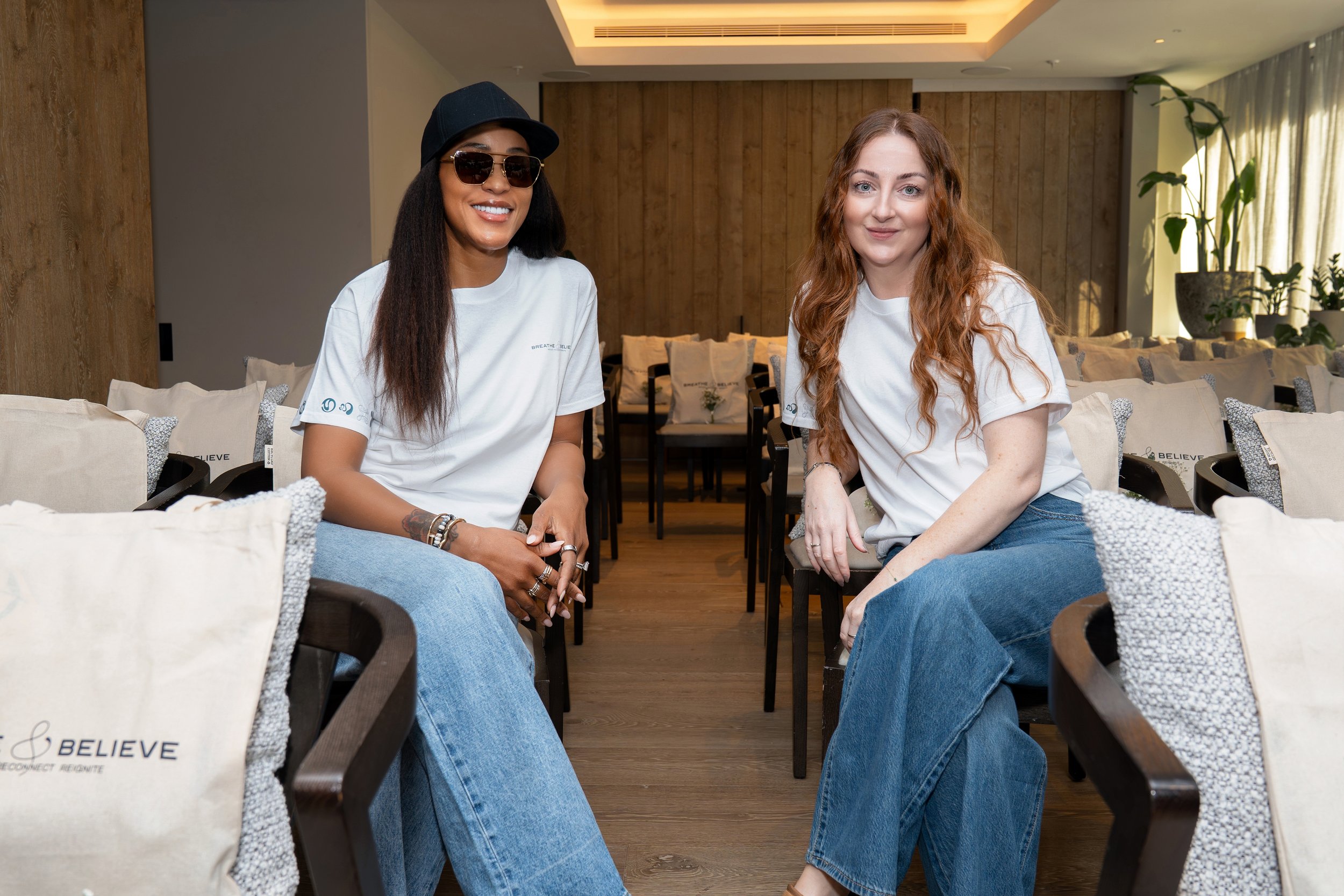Two women sitting in a cozy, modern lounge or waiting area. One woman has long dark hair, sunglasses, and a black hat, and is smiling at the camera. The other woman has long wavy red hair and is also smiling, with a relaxed posture. They are wearing casual white t-shirts and blue jeans. The background features wooden wall paneling, large windows with curtains, indoor plants, and beige pillows with the word "BELIEVE" printed on them.