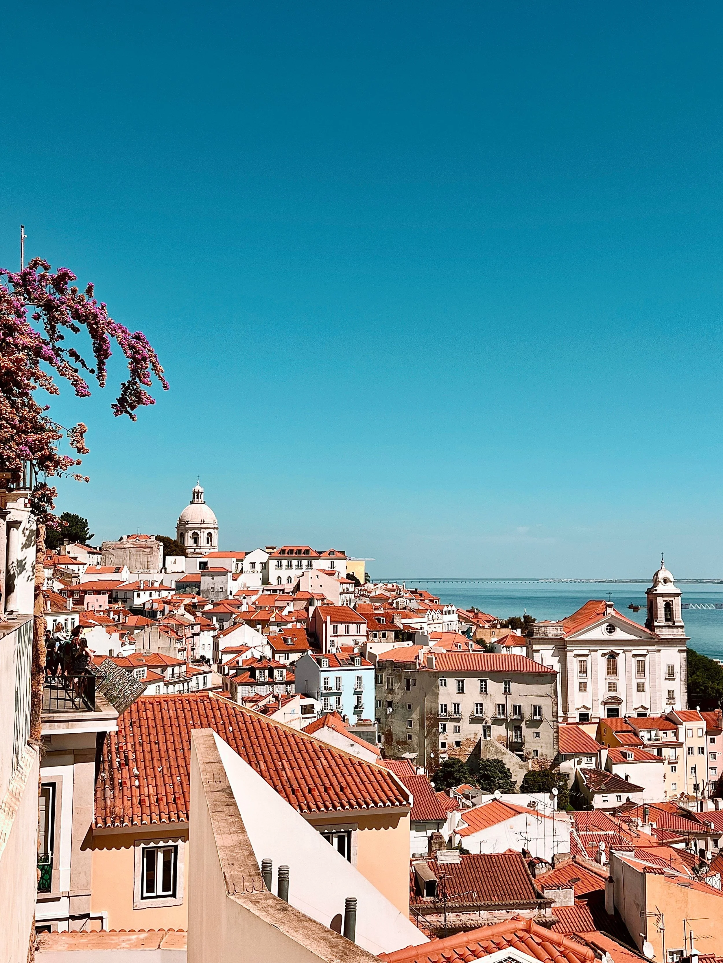 View of a city with orange-tiled rooftops, church domes, and bell towers under a clear blue sky by the water.