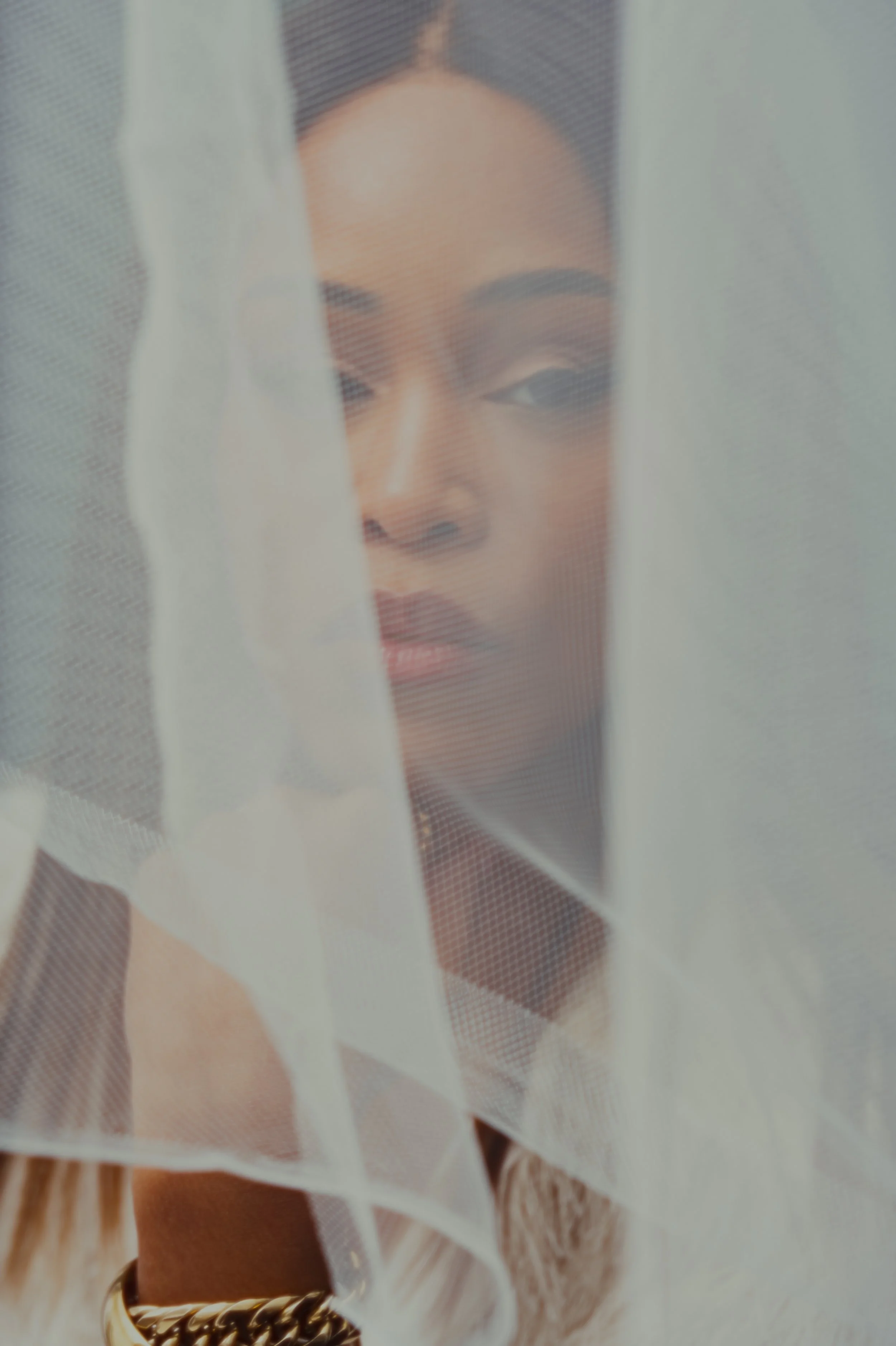 A woman with a dark complexion and dark hair behind a sheer curtain, looking at the camera.