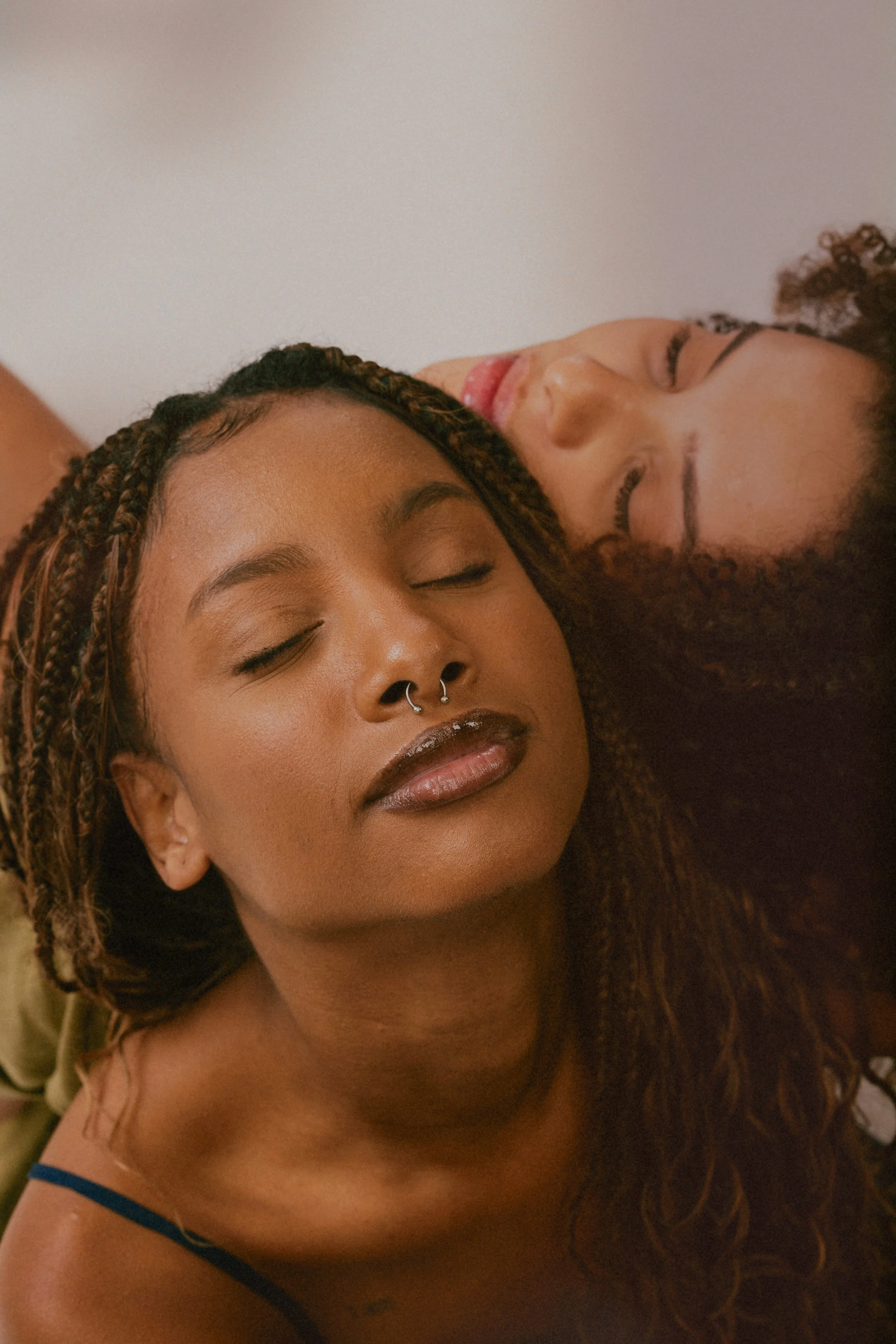Two women with closed eyes, relaxed expressions, lying close together, one with braided hair and a nose ring, the other with curly hair, against a plain background.