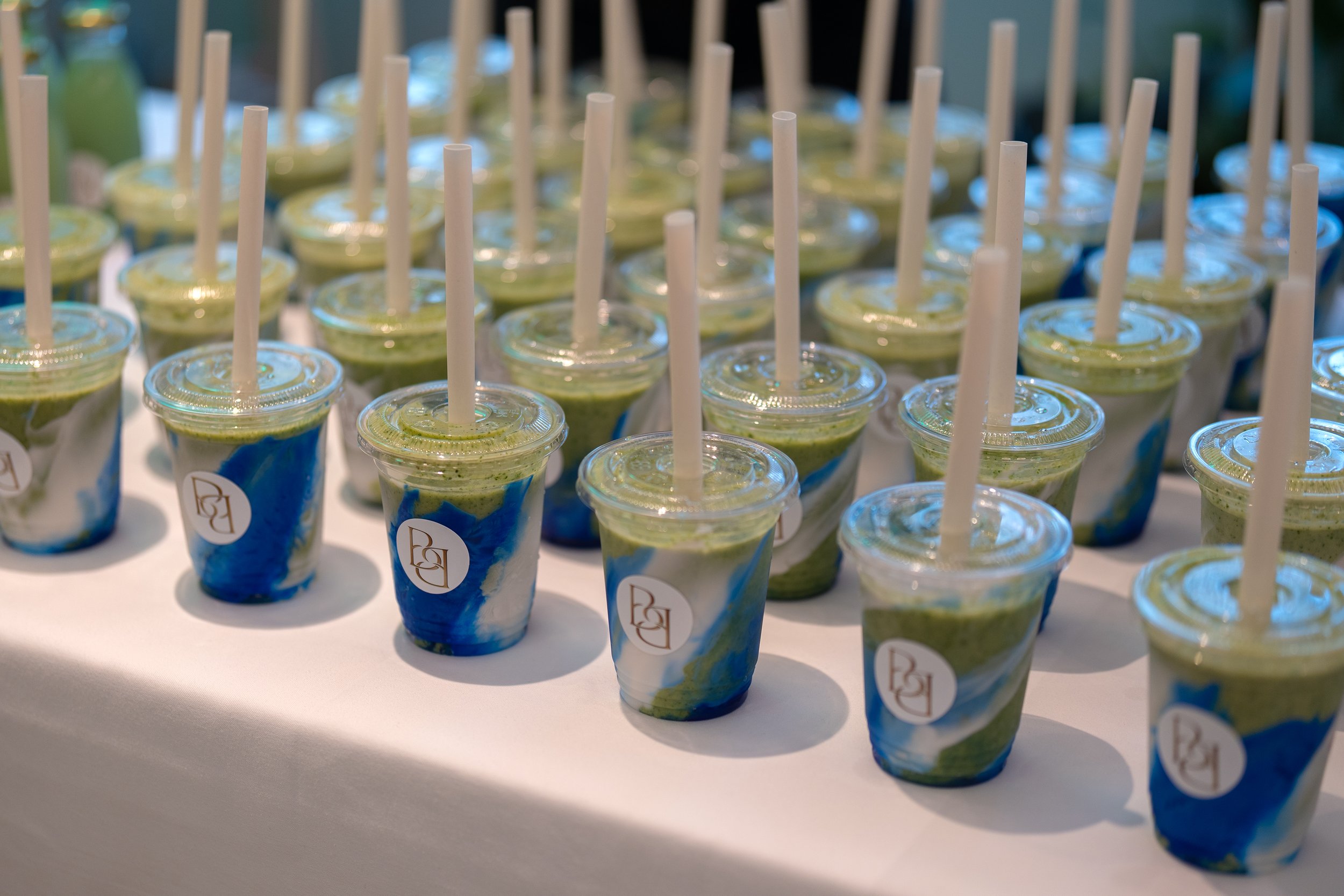 Multiple cups of green and blue smoothies with white straws on a white surface, with a blurred background.