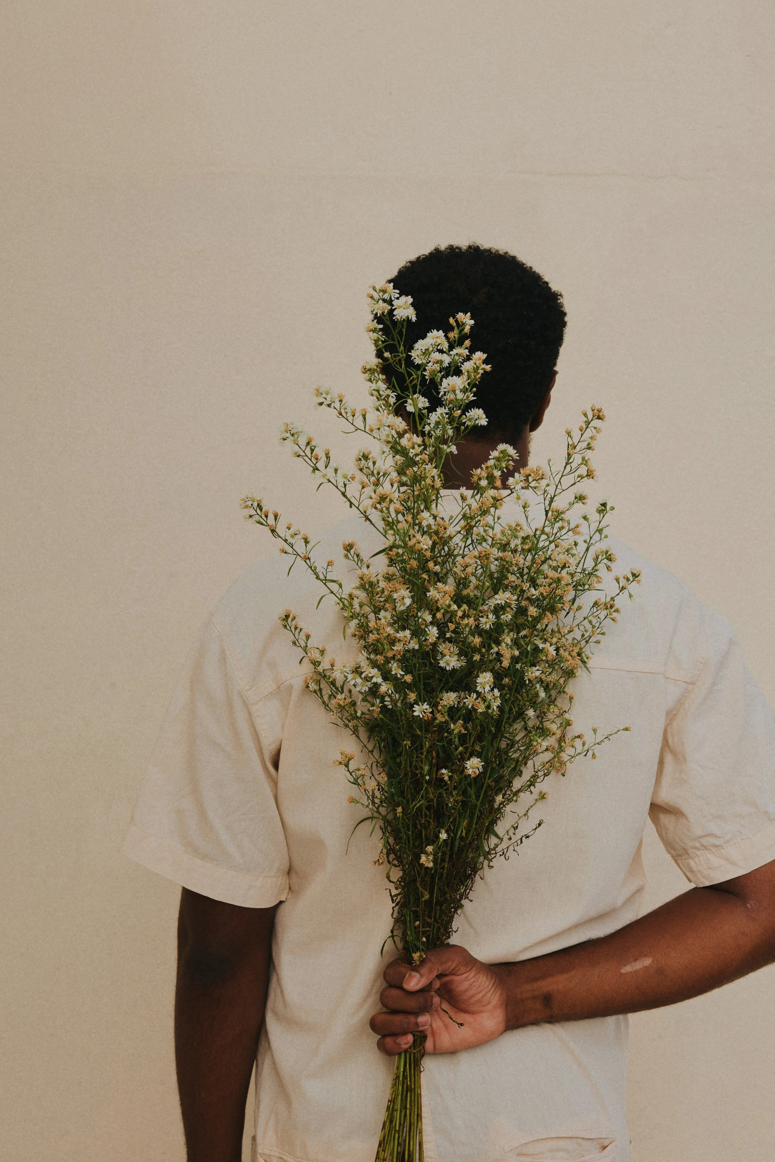 A person with dark curly hair holds a bunch of small white flowers behind their back, facing away from the camera, against a beige background.