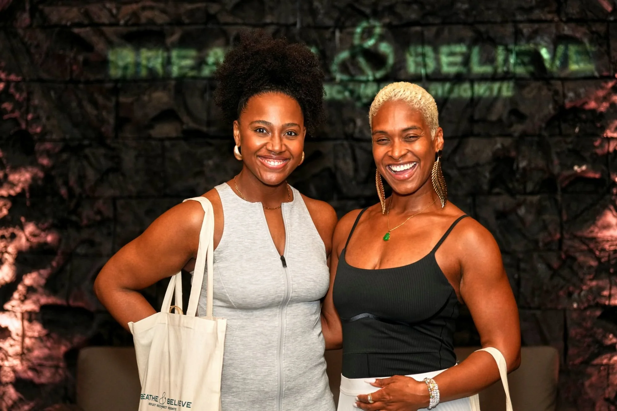Two women smiling, one with short curly black hair wearing a light gray sleeveless top, and the other with short blonde hair wearing a black tank top, standing in front of a dark textured wall with green and pink lighting accents.