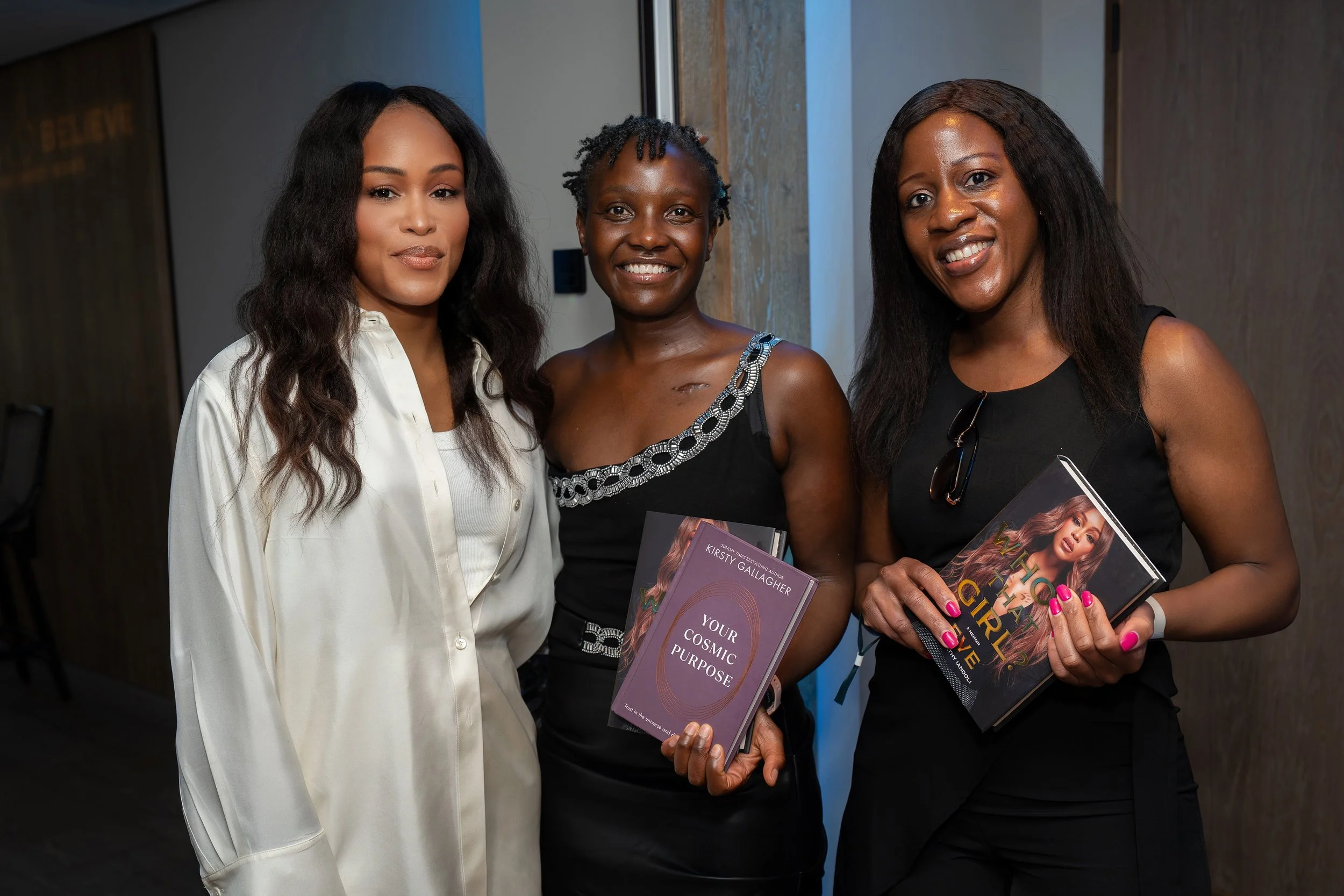 Three women standing together indoors holding books and smiling at the camera. One woman on the left wears a white shirt, the middle woman wears a black dress with silver detailing, and the woman on the right wears a black top with sunglasses hanging from it. The background includes a wooden wall and a blue-lit area.
