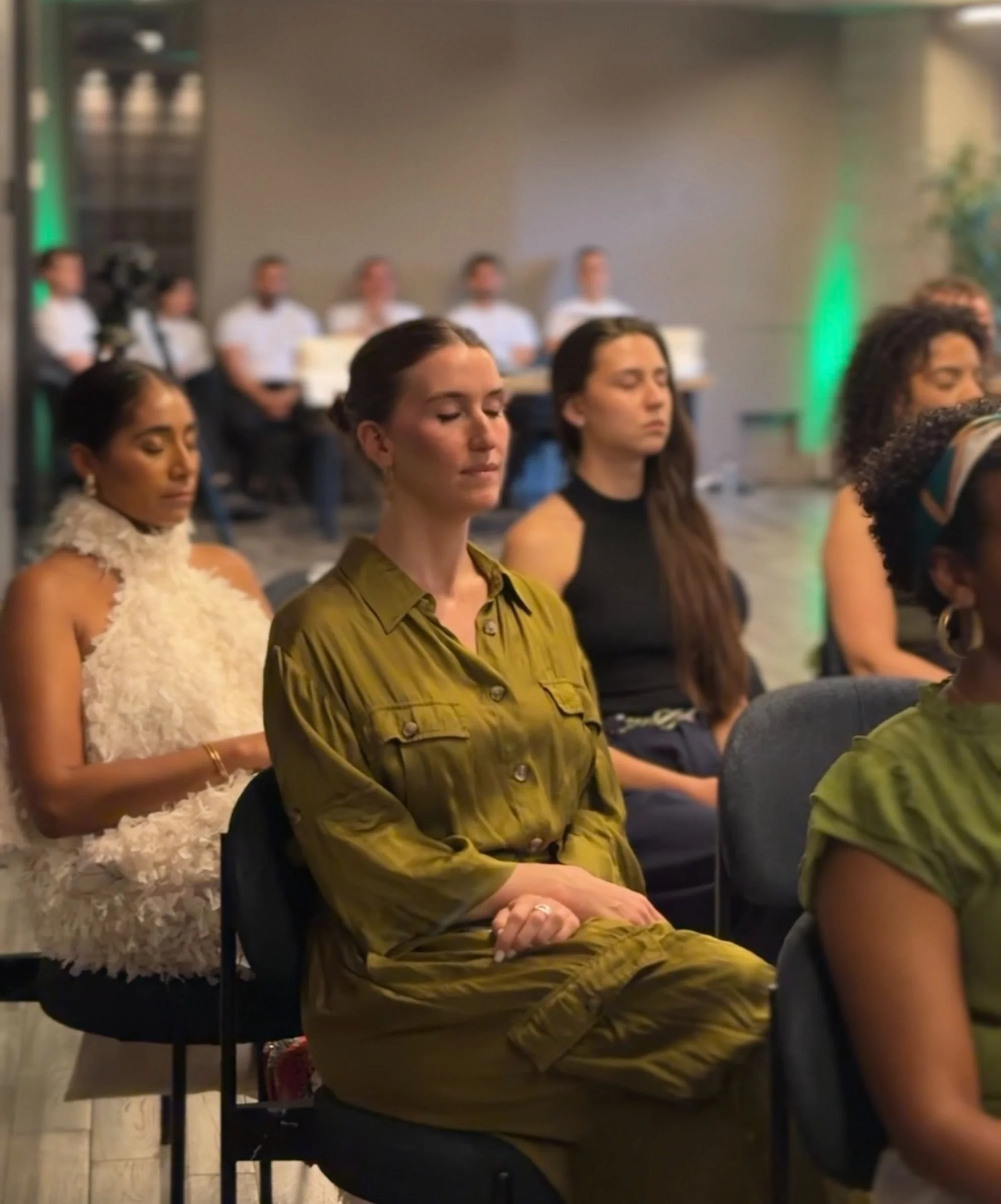 People sitting with eyes closed, possibly meditating or attending a mindfulness session, in a room with gray walls and some green lighting.