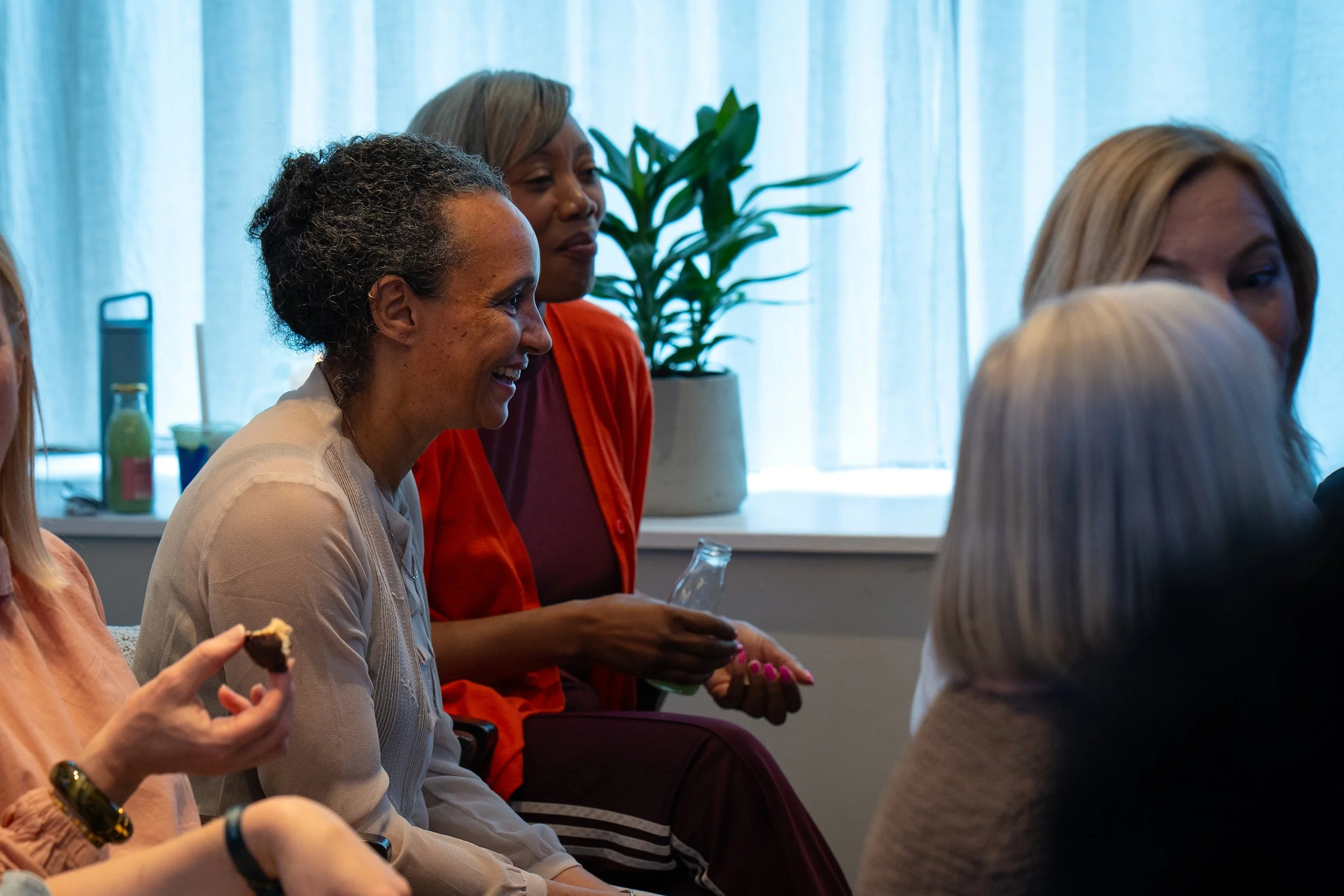 Group of women sitting indoors, engaging in a conversation or meeting, with a window and a plant in the background.