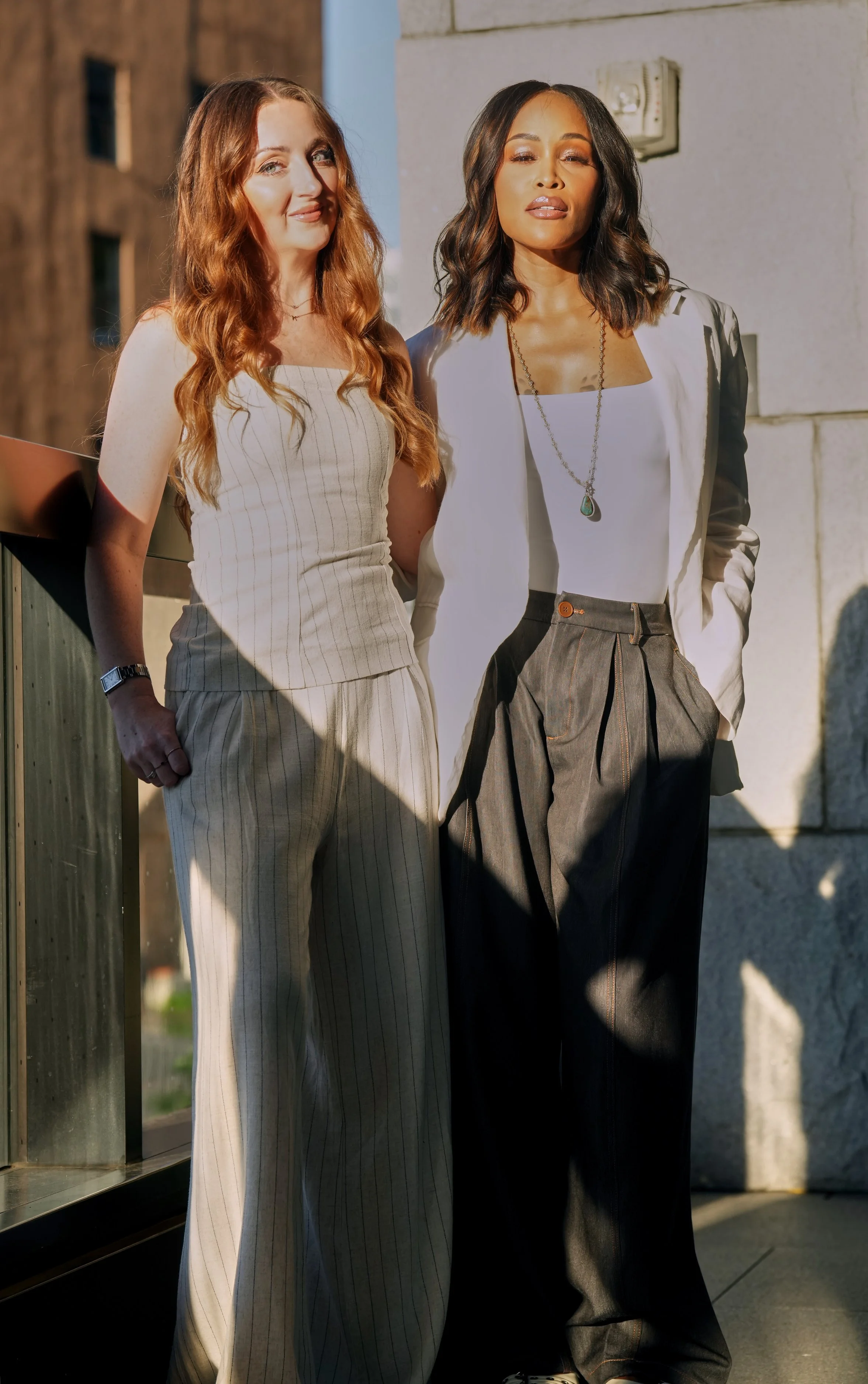 Two women standing outdoors beside a wall, dressed in stylish clothing, with natural sunlight illuminating their faces.
