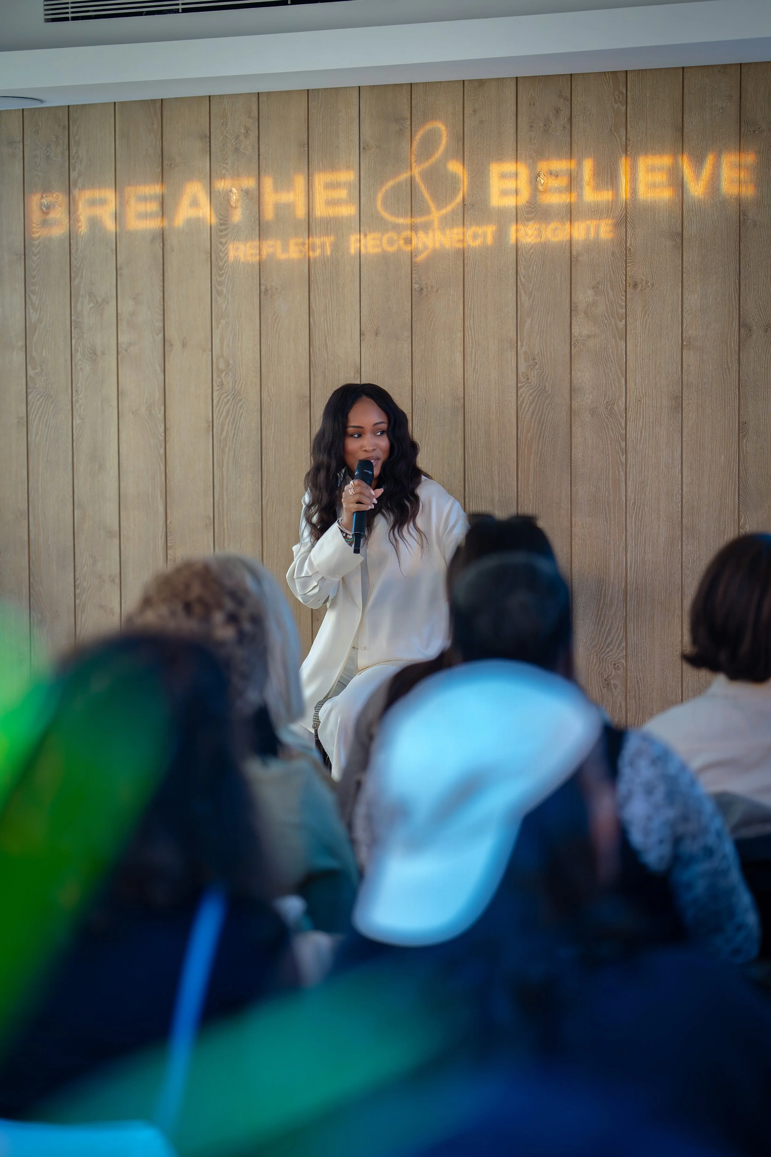 A woman with long dark hair, wearing a white blazer, speaking into a microphone during a presentation or speech, in front of a wooden-paneled wall with illuminated words 'Breathe & Believe' and the phrase 'Reflect, Reconnect, Radiate'