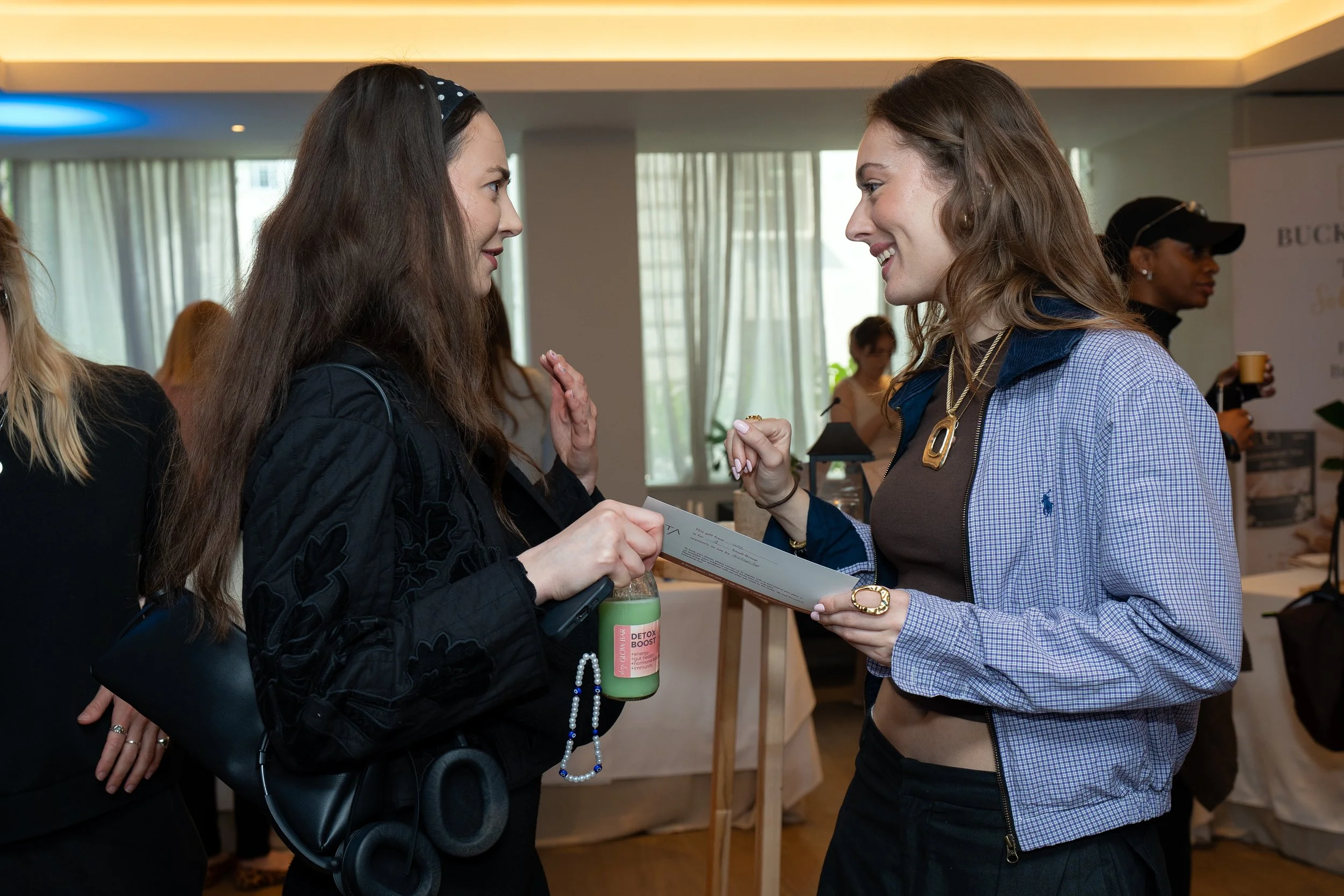 Two women are engaged in a conversation indoors, smiling, with one woman holding a certificate or document, and the other woman holding a green bottle. There are other people and a BUCK logo in the background.