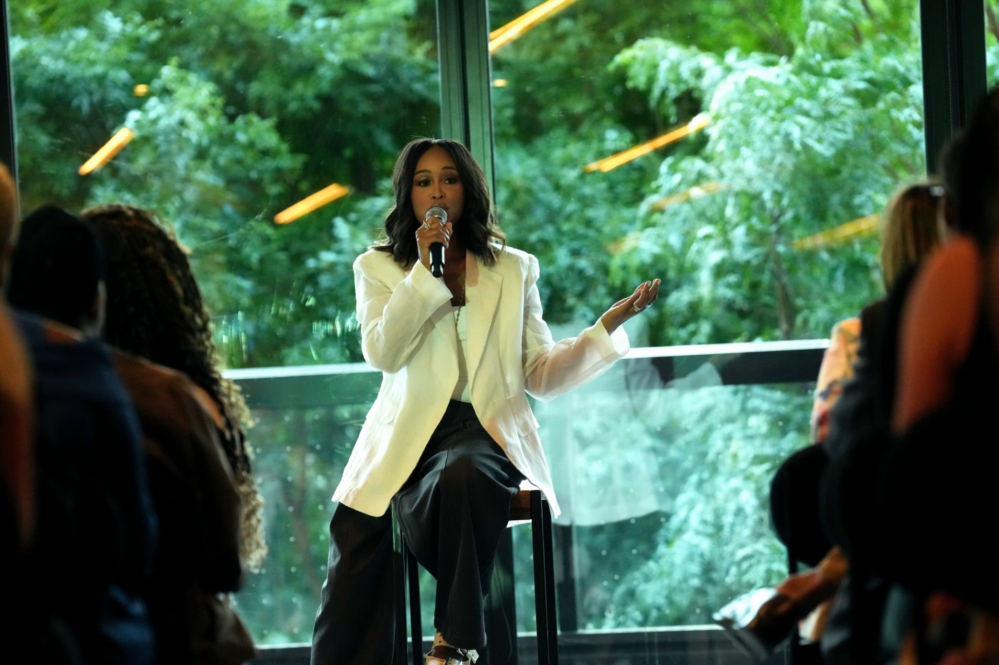 A woman in a white blazer and dark pants presenting with a microphone in front of an audience in a room with large windows showing green trees outside.