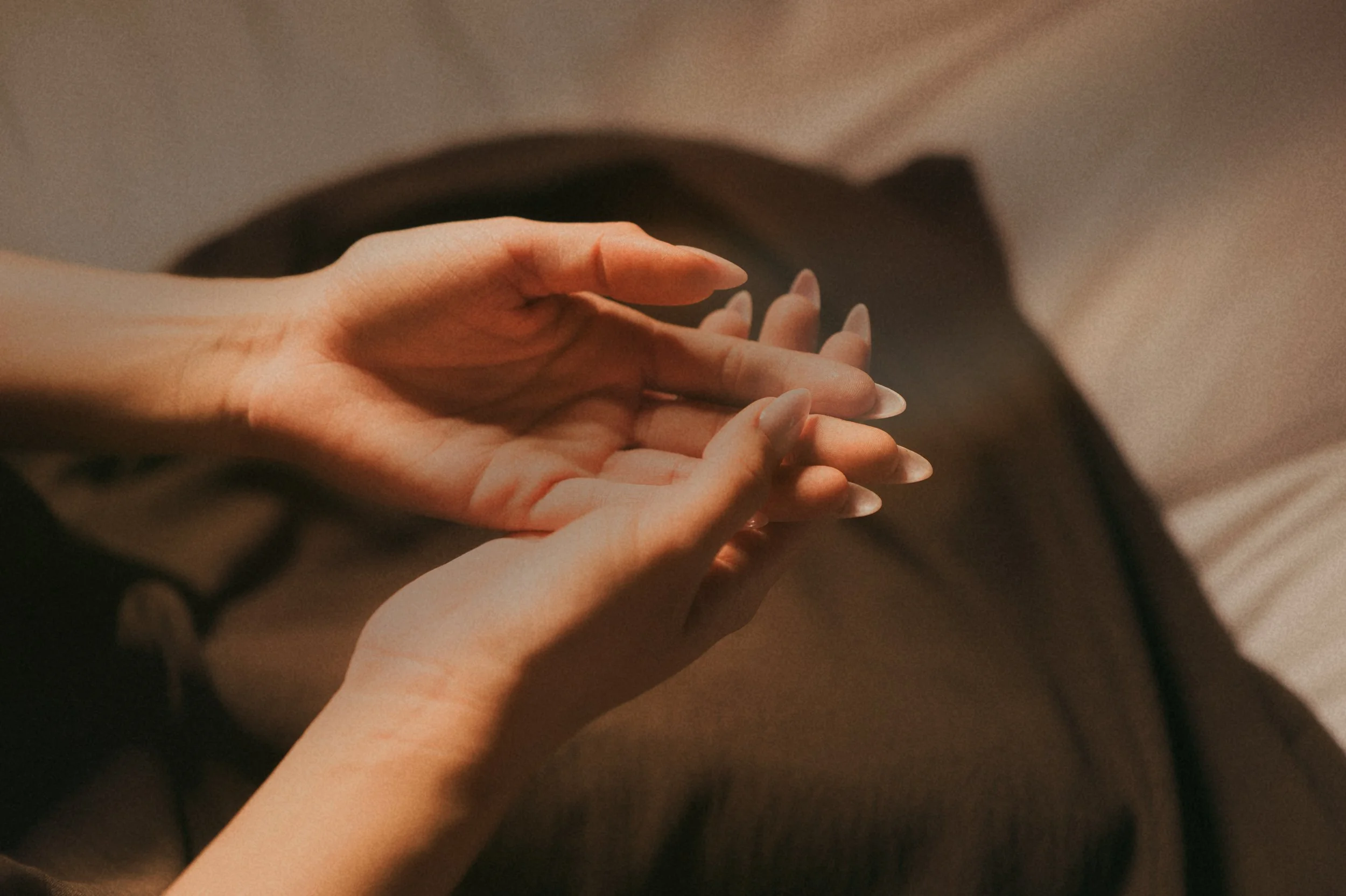Close-up of two hands with light skin and long nails gently holding each other, resting on a dark surface.
