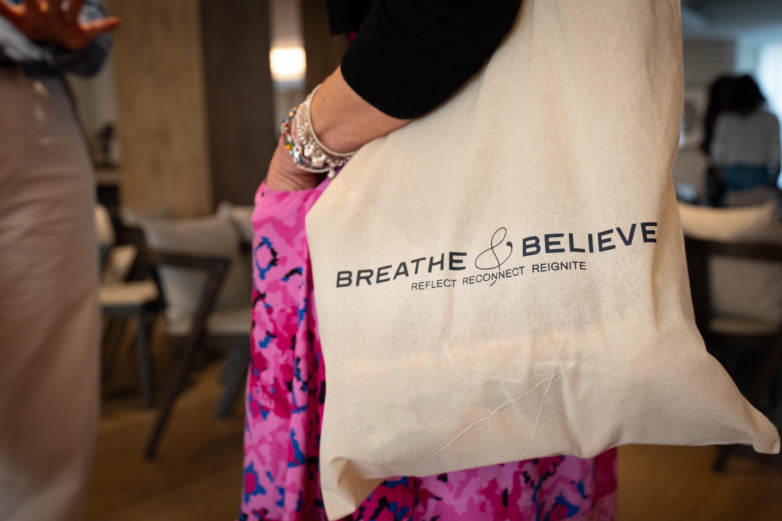 Person wearing a white tote bag with the words "BREATHE & BELIEVE" and the phrases "REFLECT RECONNECT REIGNITE" printed on it, standing near other people in an indoor setting.