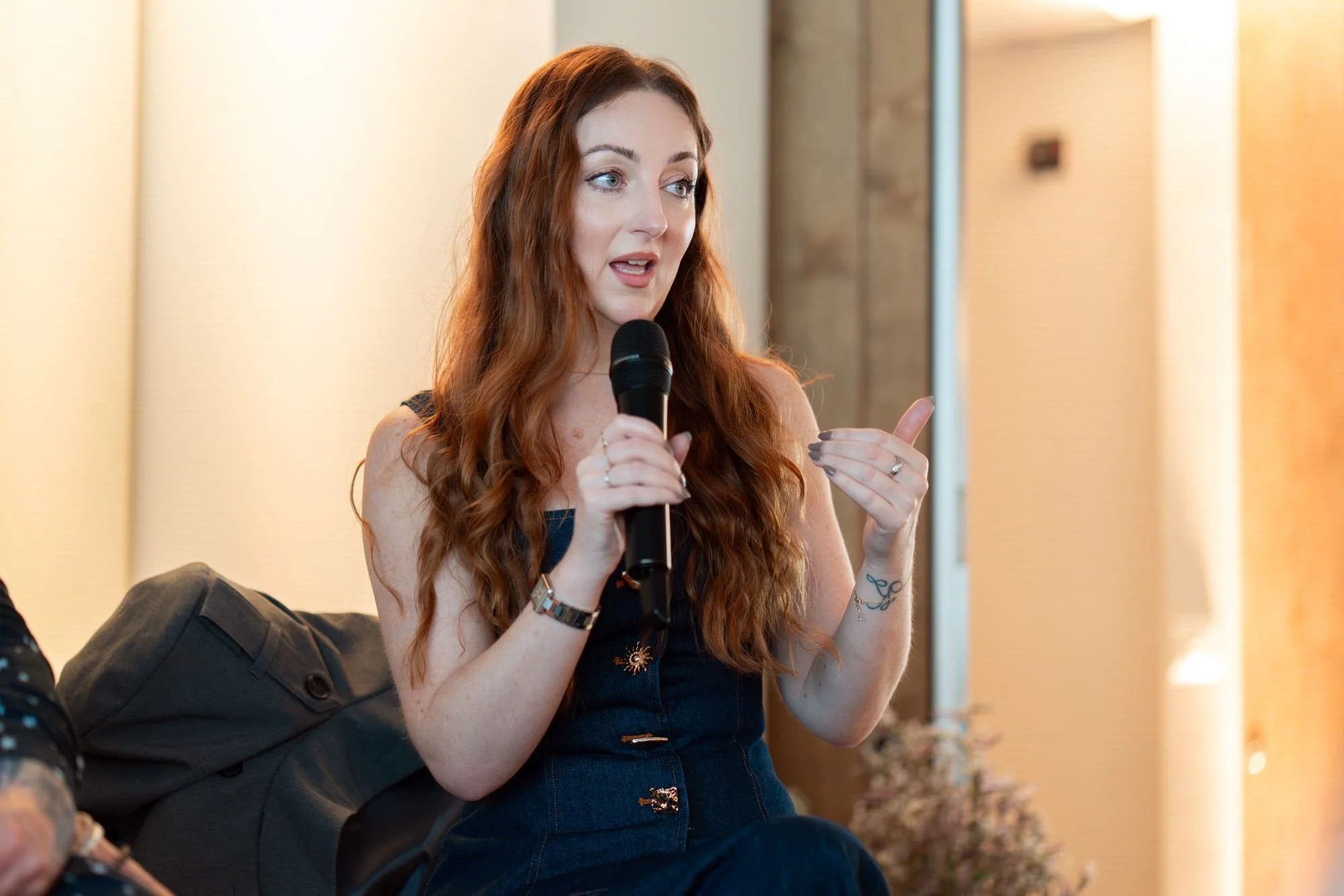 A woman with long, curly red hair speaking into a microphone during an indoor event.