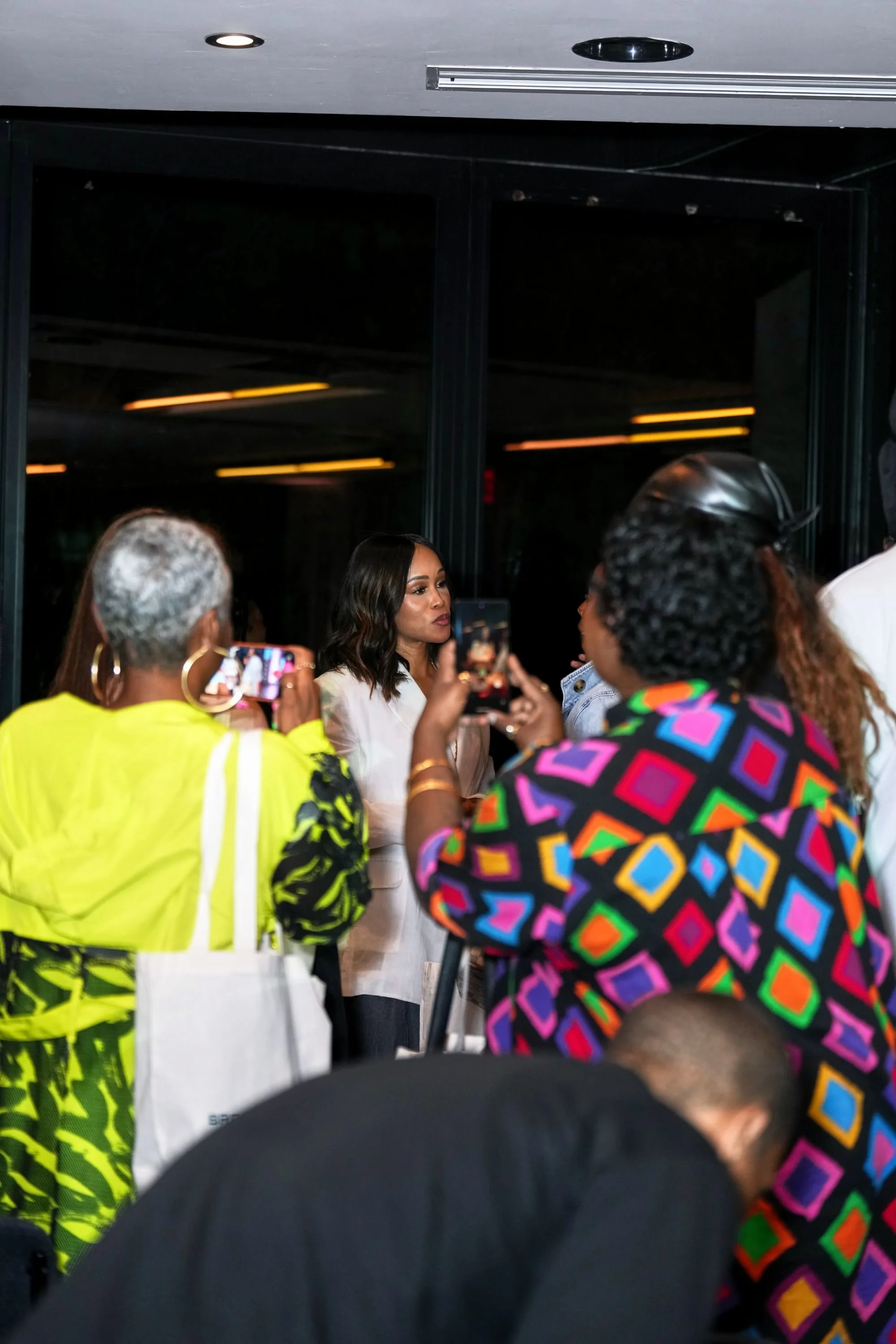 A group of women gathered, with one woman taking a photo of another woman who is speaking. The women are wearing colorful and black clothing, engaged in conversation in an indoor setting.