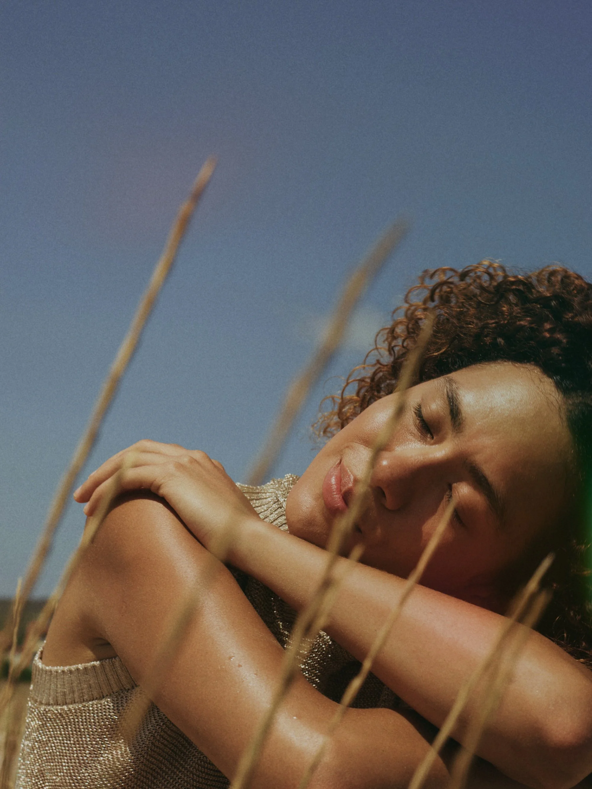A woman with curly hair and closed eyes resting her head on her crossed arms outdoors, with a clear blue sky and tall grass in the foreground.