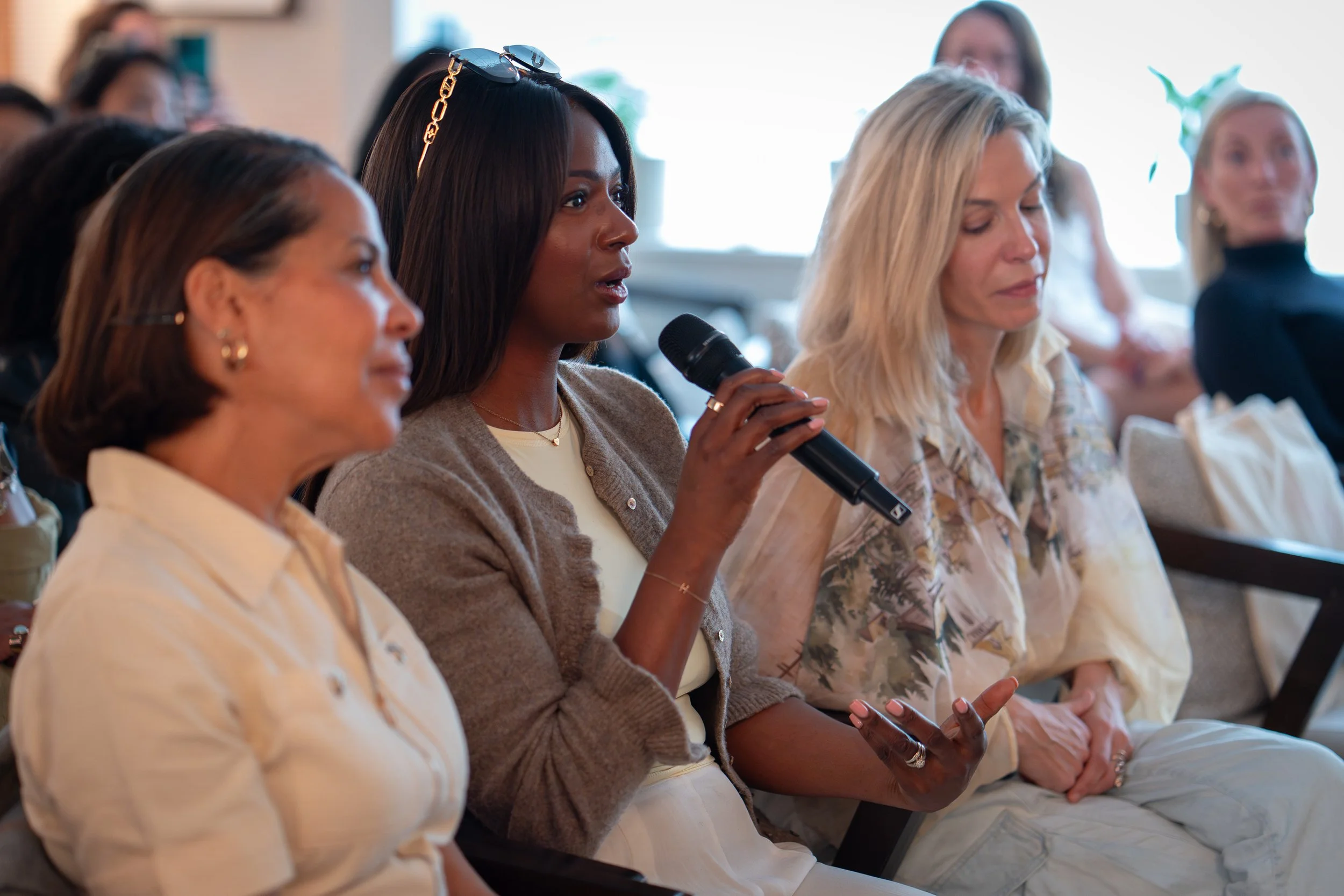A woman with dark hair holding a microphone, listening attentively during a discussion at a conference or meeting with other women present.