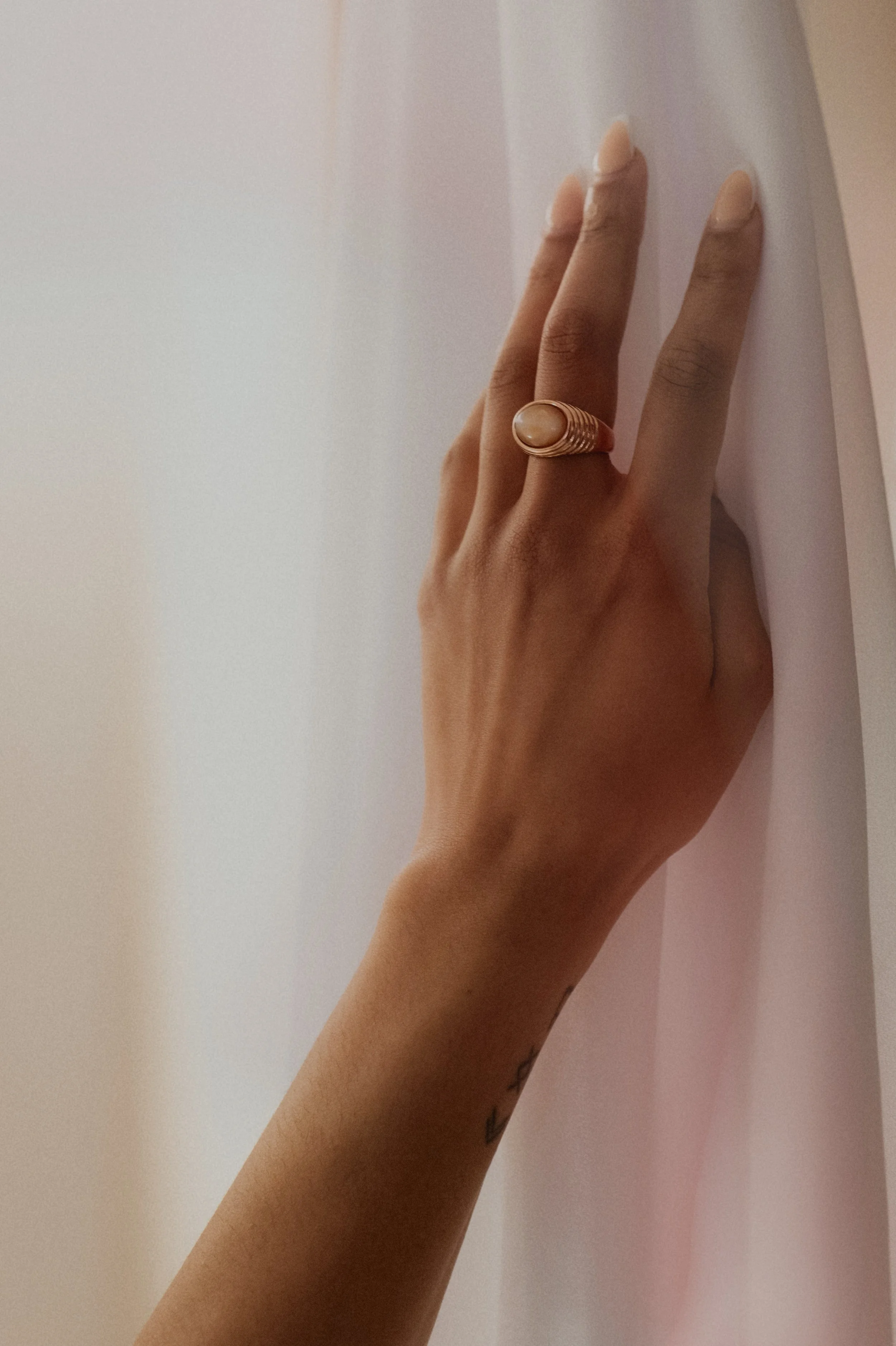 A person's hand with light pink nails and a large gold ring touching a white curtain.