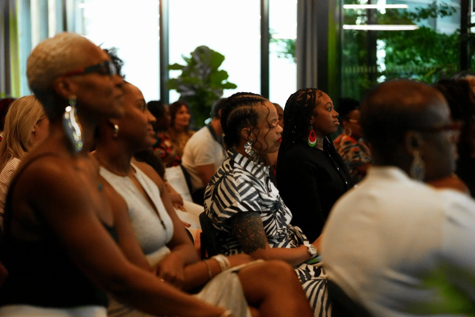 A group of women seated attentively at a conference or seminar, in a brightly lit room with large windows, greenery visible outside.