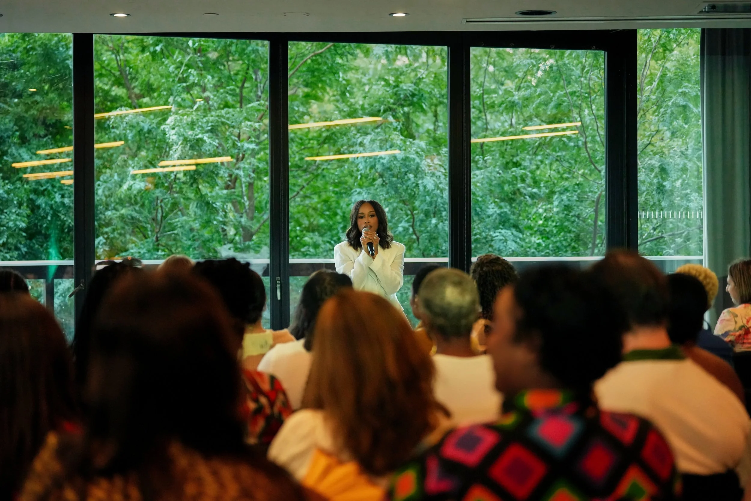 A woman in a white blazer is speaking into a microphone at an indoor event with large windows revealing green trees outside. The audience is seated and facing her.