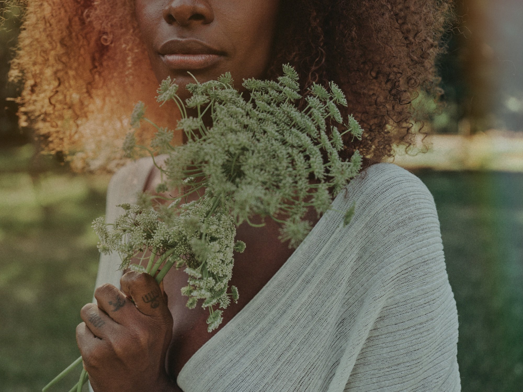 A woman with curly hair holding a bunch of wildflowers close to her face outdoors.