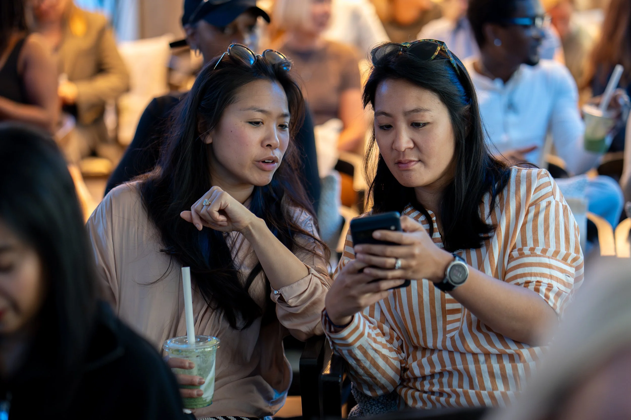 Two women are looking at a smartphone together at a crowded outdoor event, with people in the background. One woman is holding a green and white beverage in a clear plastic cup with a straw.