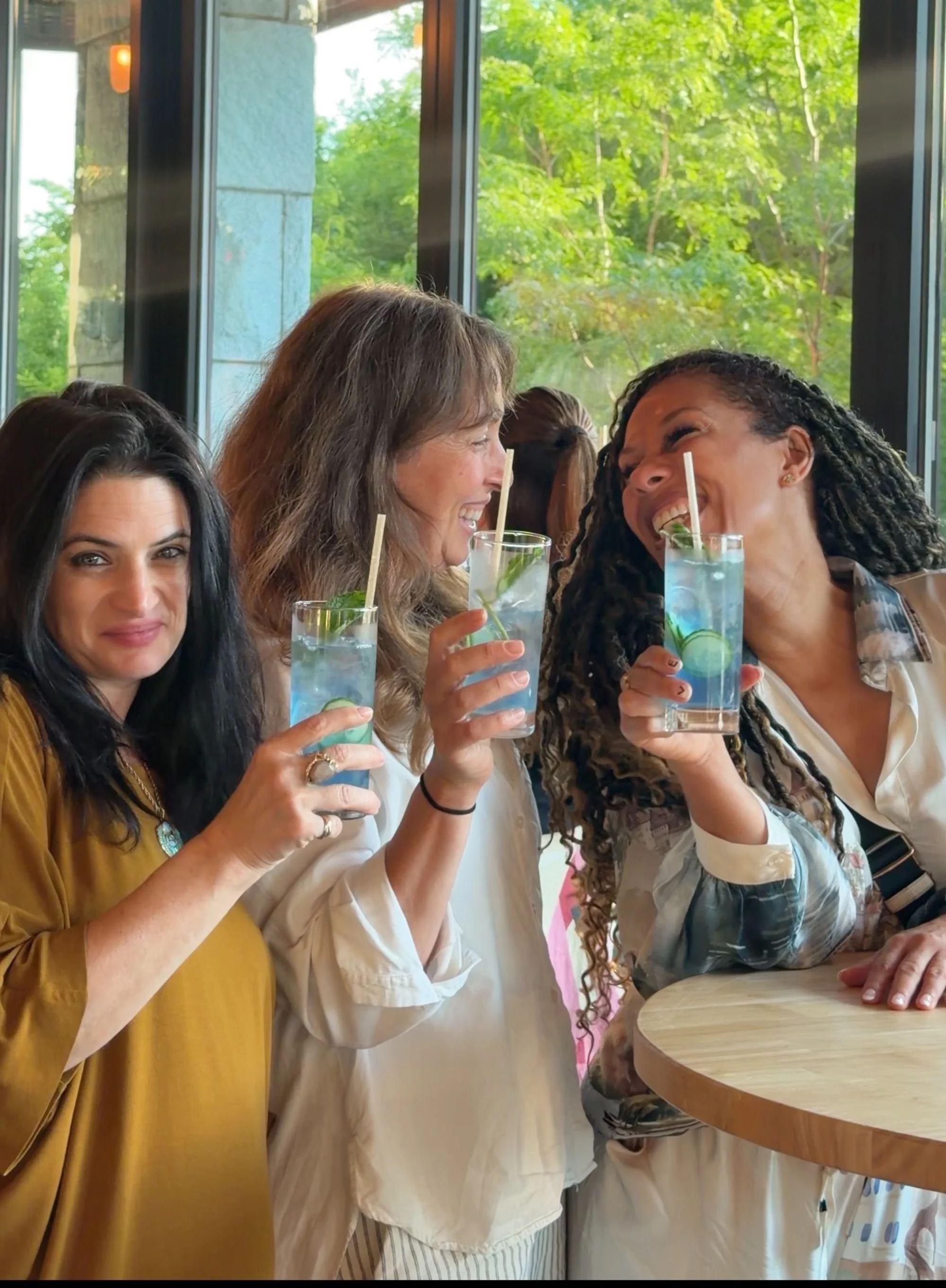 Three women smiling and laughing while holding cocktails in a restaurant with large windows and green trees outside.