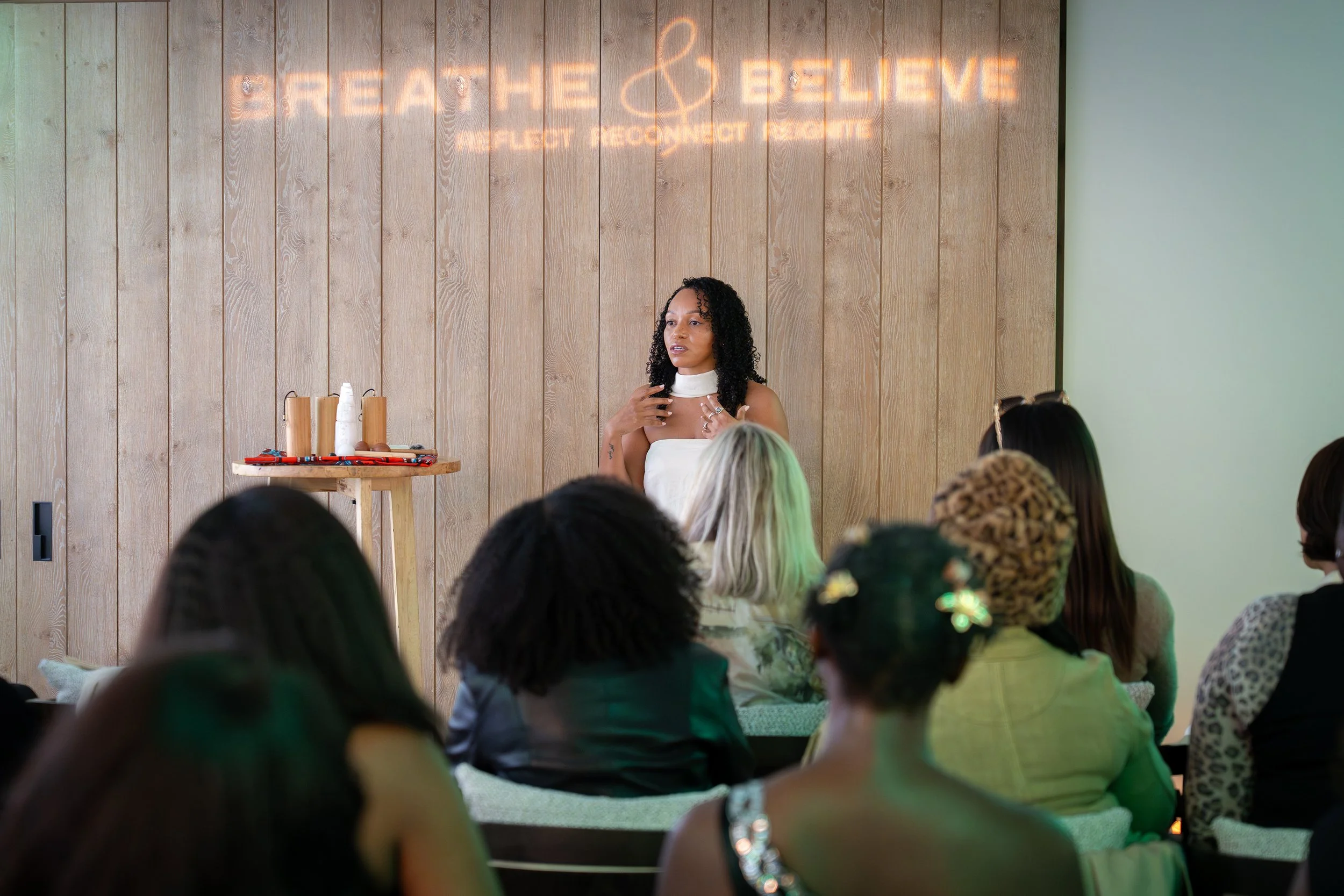 A woman speaking to an audience during a presentation in a room with wooden wall panels and a neon sign that reads 'PEACE & BELIEVE'.