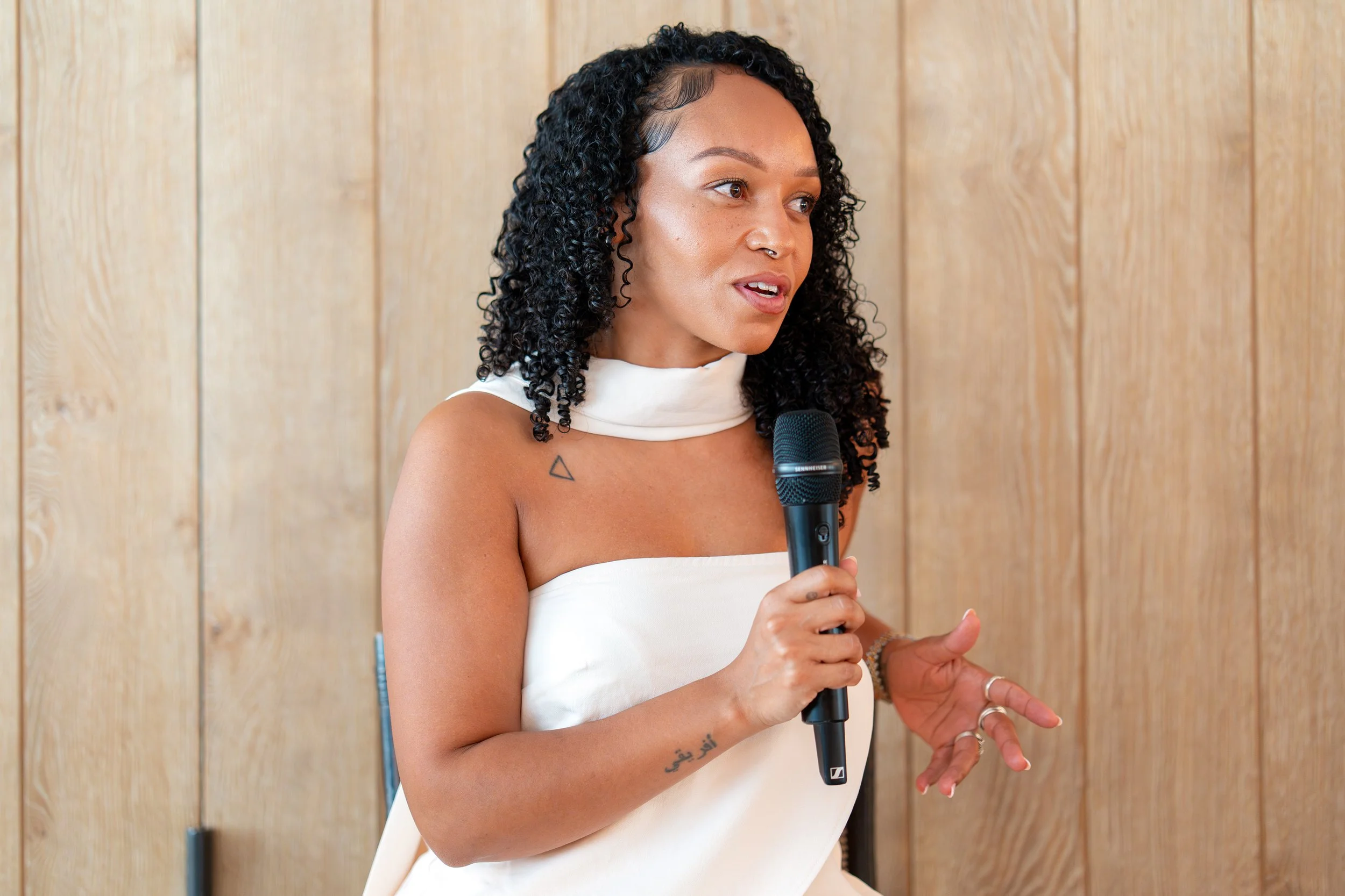 A woman with curly black hair and tattoos on her arm is speaking into a microphone, standing against a wooden wall.