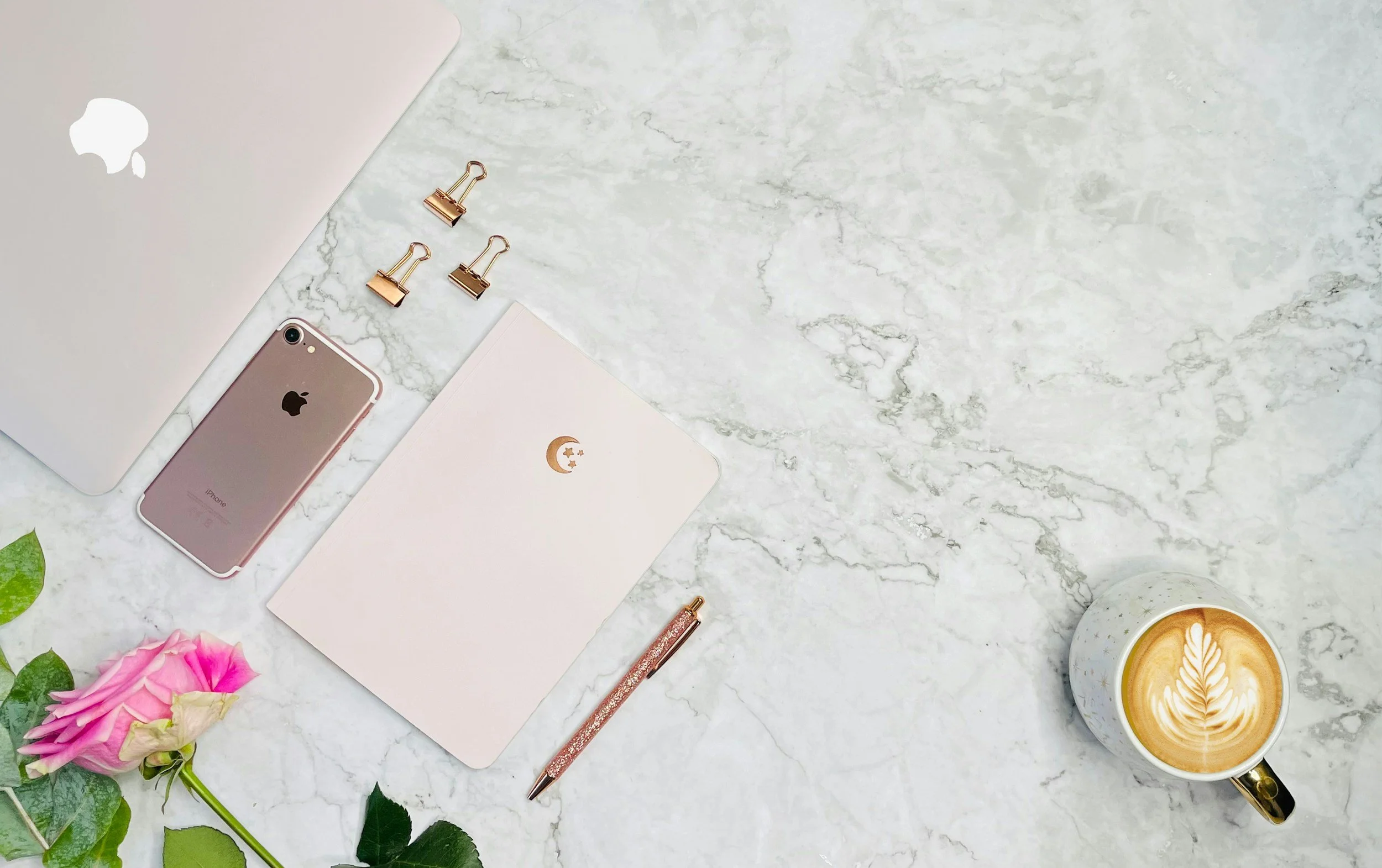 A flat lay of a marble surface with a closed silver MacBook laptop, a rose gold iPhone, a pink notebook with a gold crescent moon and star design, three gold binder clips, a pink and gold glitter pen, a cup of coffee with latte art, and pink roses with green leaves.