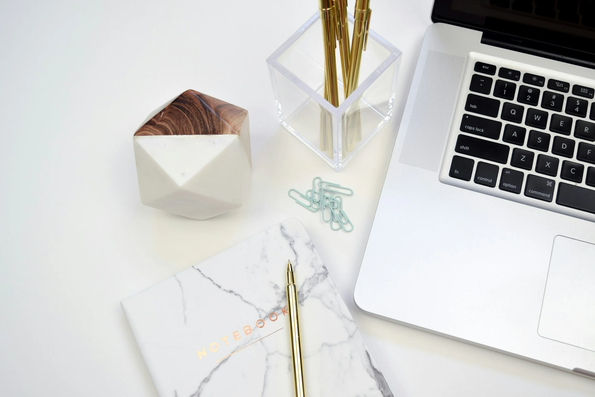 An organized desk workspace with a macbook laptop, a clear acrylic pen holder with gold pens, a marble-patterned contact paper, a gold pen, a small notebook labeled 'NOTEBOOK' with a marble cover, paper clips, and a geometric wooden block.